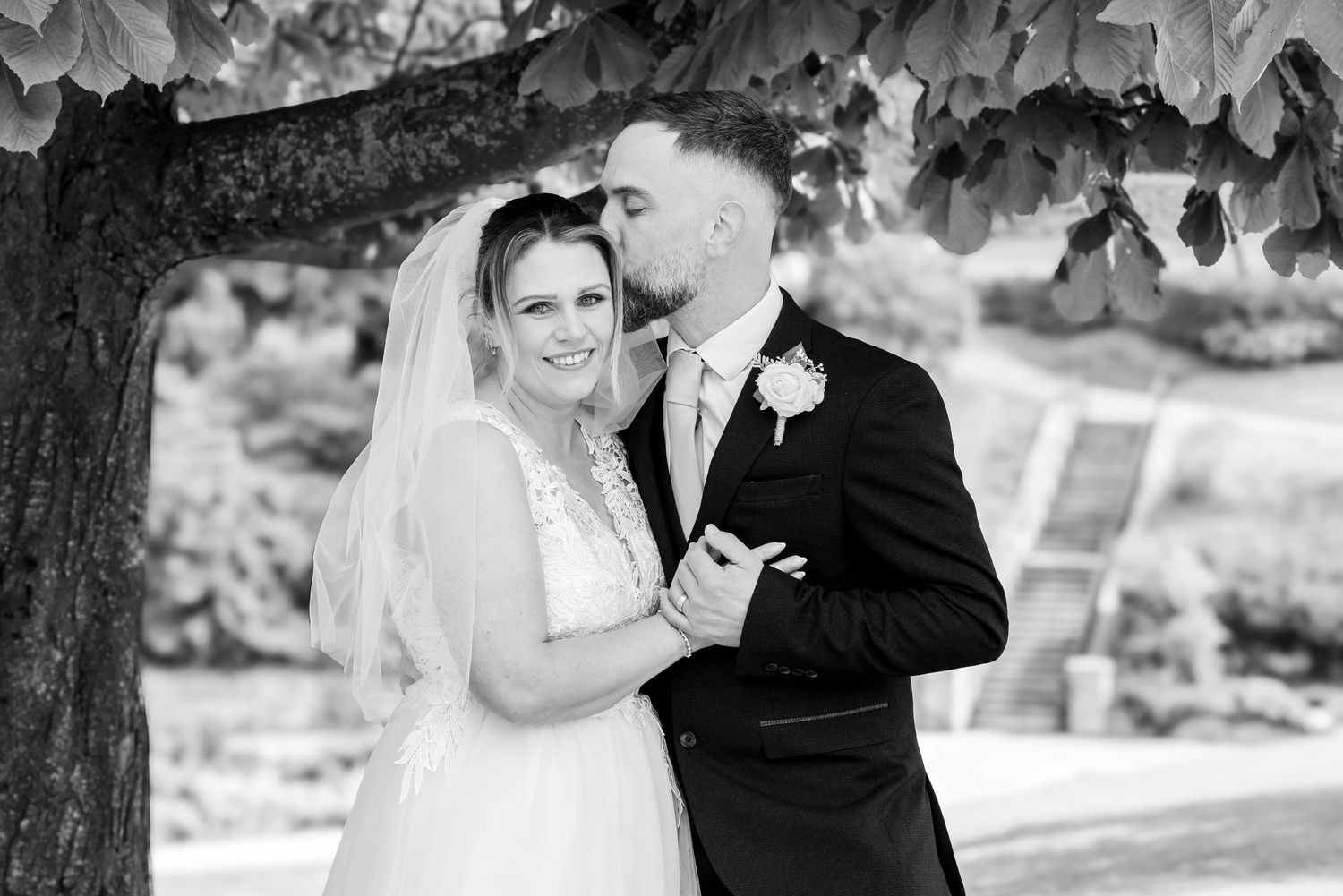 Black and white wedding photo of bride and groom embracing under a tree, groom kissing bride's head.