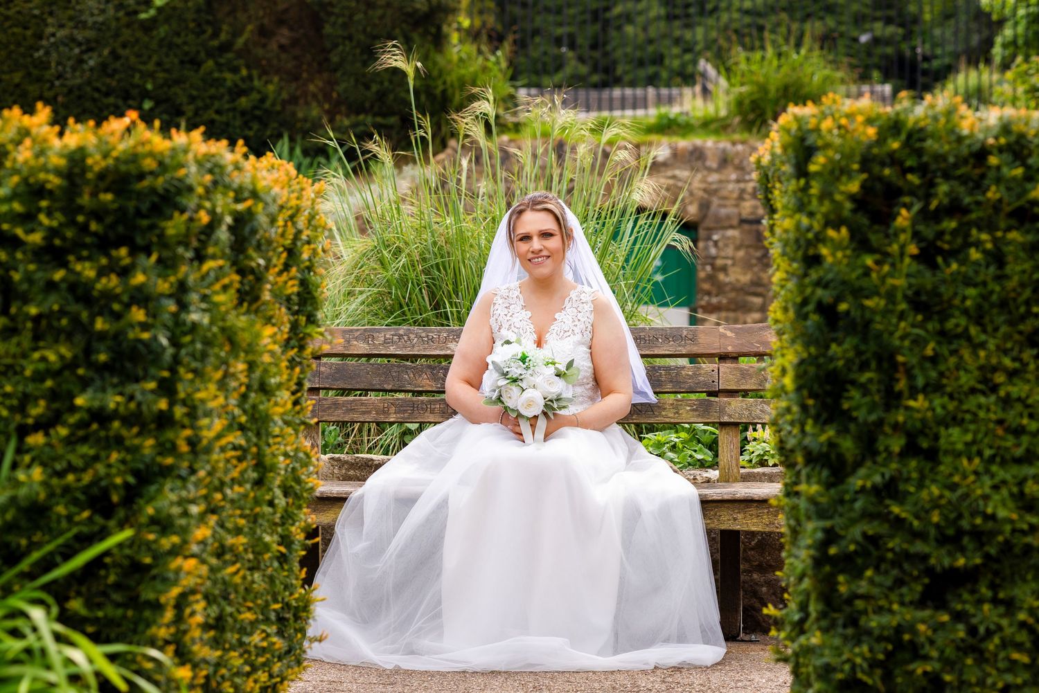 Bride in white gown and veil sits on garden bench holding bouquet, surrounded by lush green hedges.