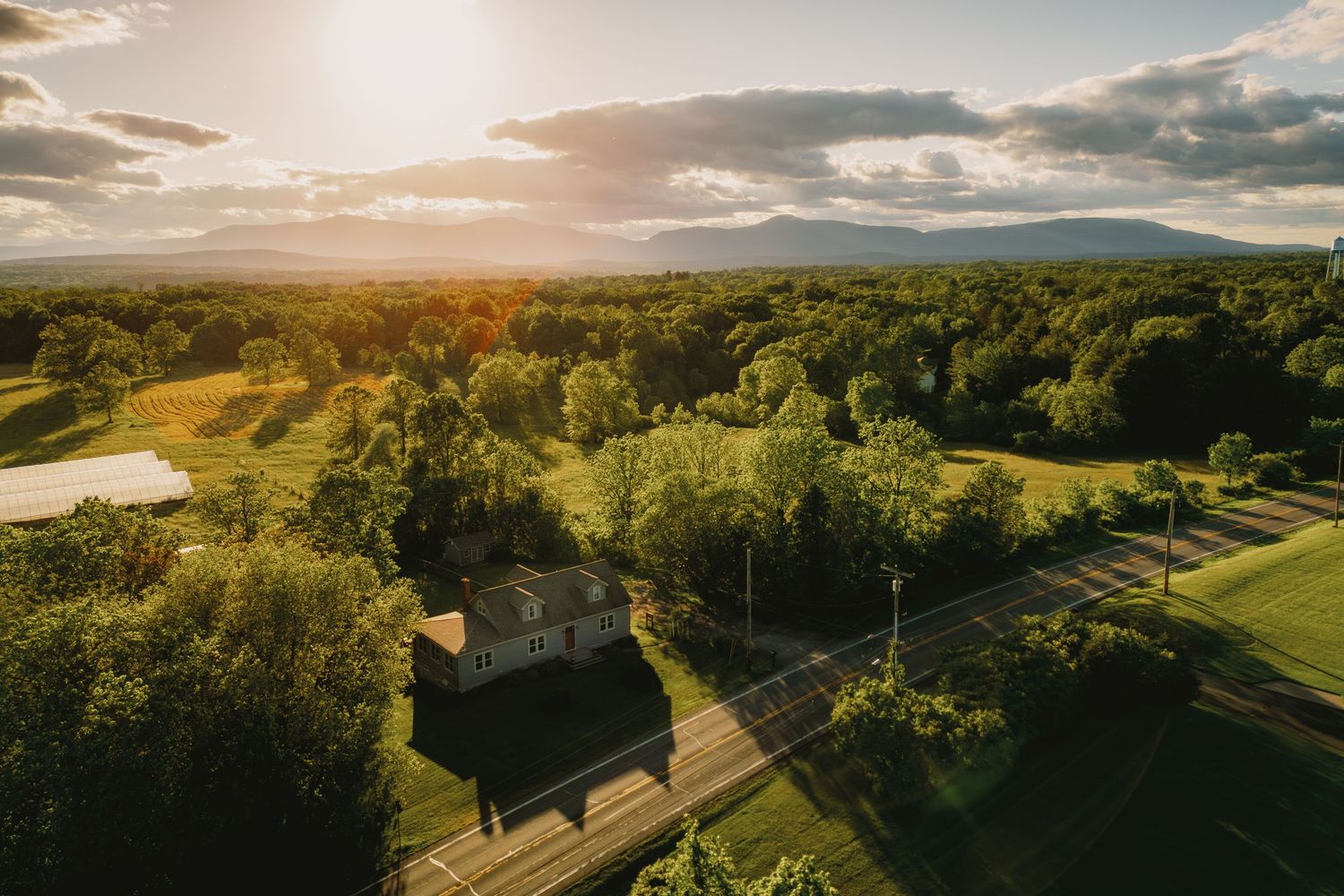Aerial sunset view of a lush green landscape with trees, a small building, winding roads, a pond, and distant mountains.