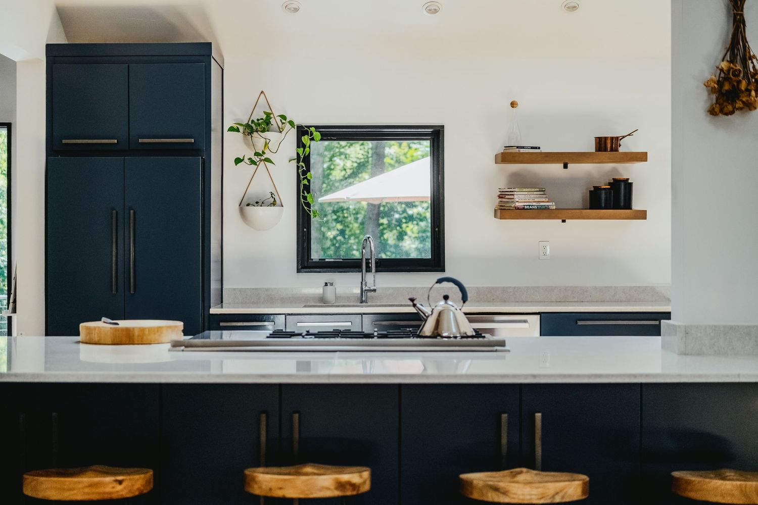 Modern kitchen features navy blue cabinets, marble countertops, open shelving and wooden barstools against white walls.
