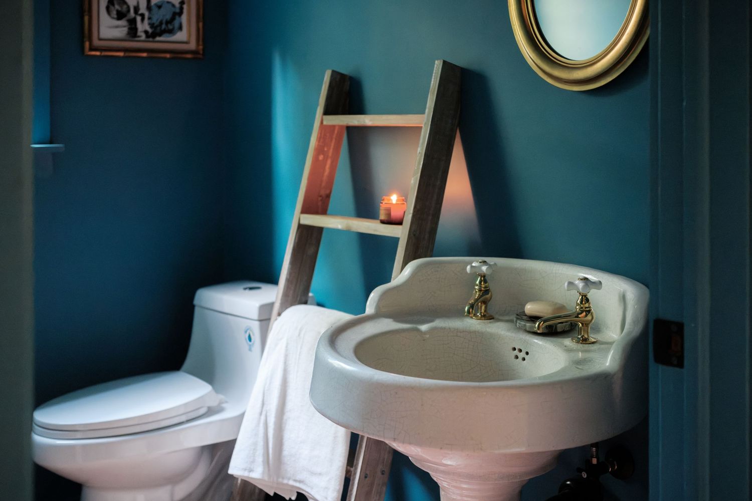 Teal bathroom with pedestal sink, brass faucets, wooden ladder shelf with candle, round gold mirror, and white toilet.