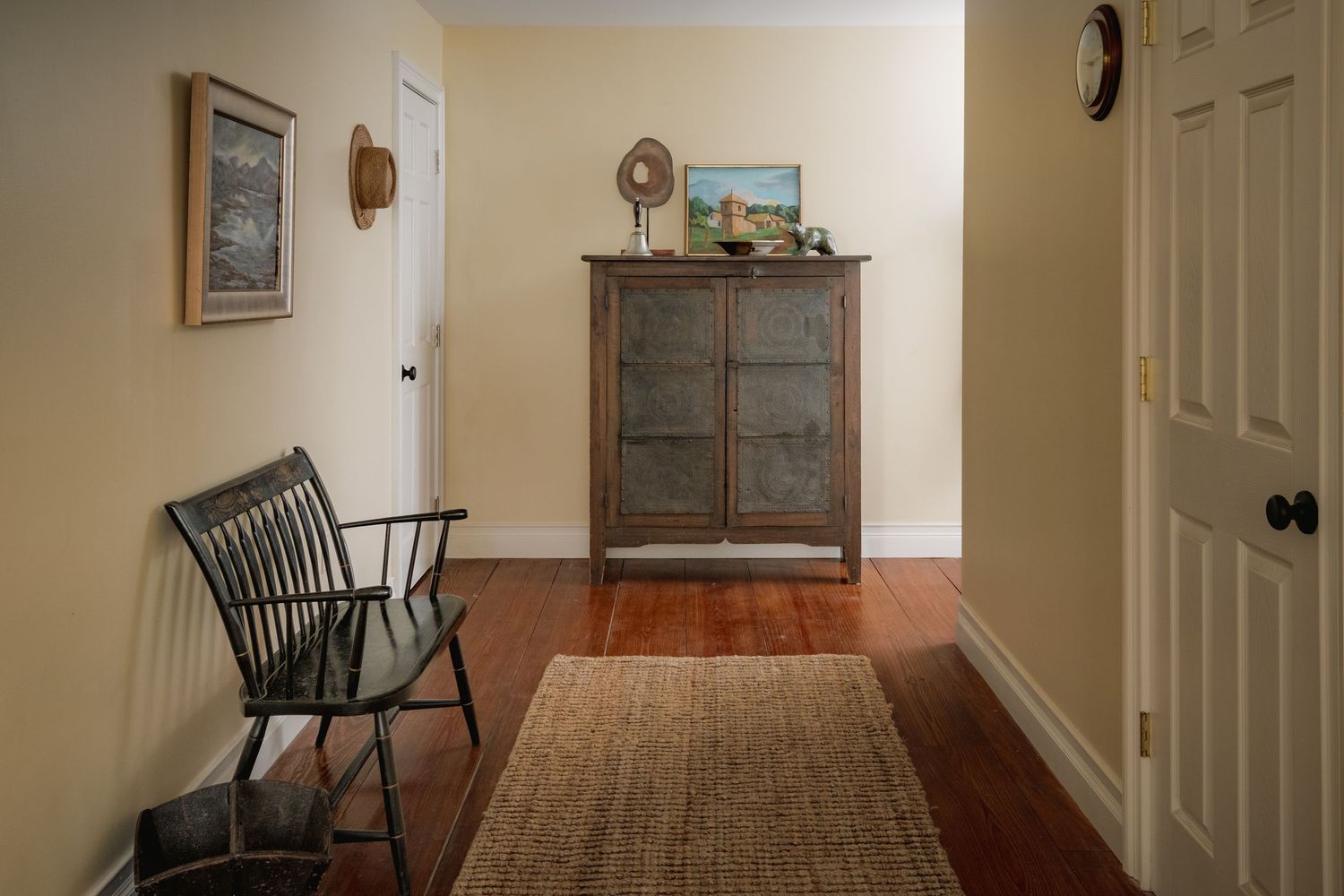 Cozy hallway with dark hardwood floors, a wooden bench, antique cabinet, framed art, and a woven rug near a white door.