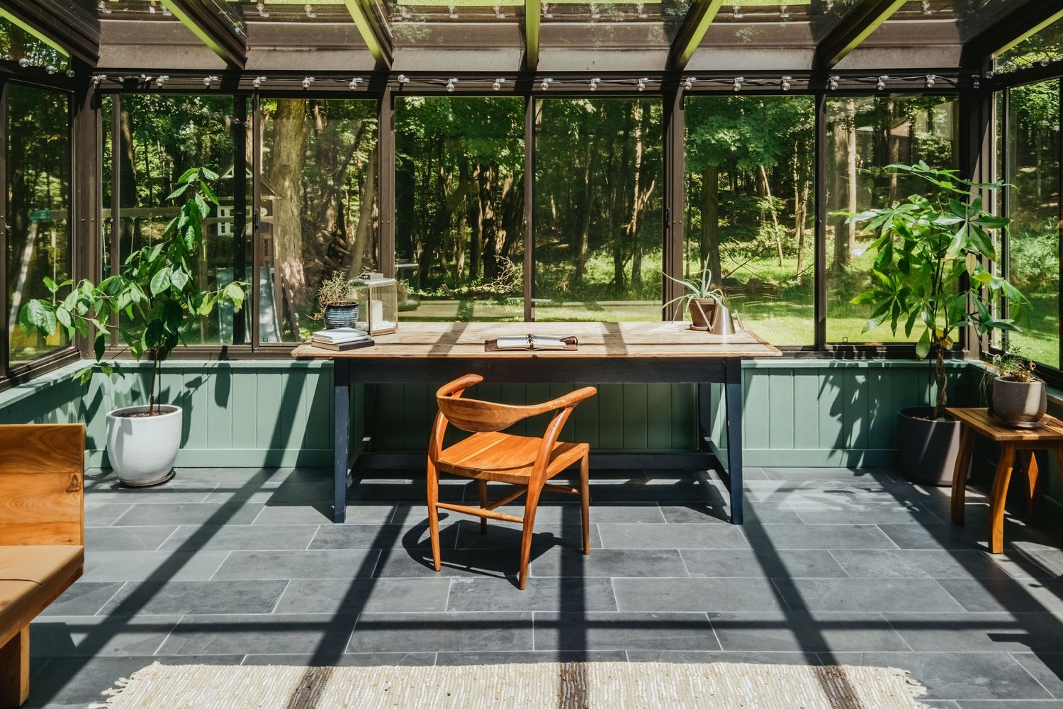 Modern office space with glass walls overlooking lush green forest, featuring wooden furniture and gray floor tiles.