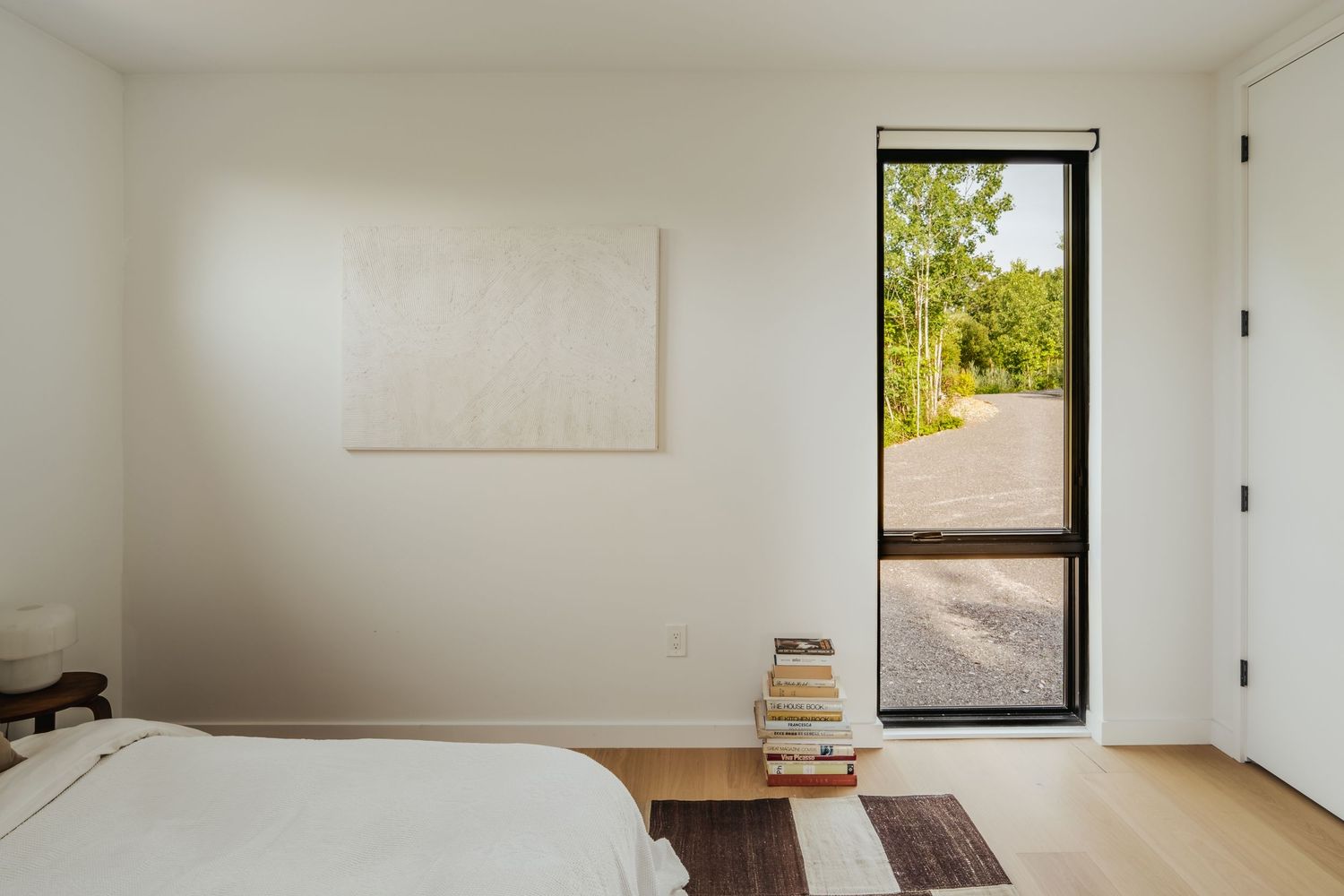 Minimalist bedroom with tall door window and stacked books on floor.