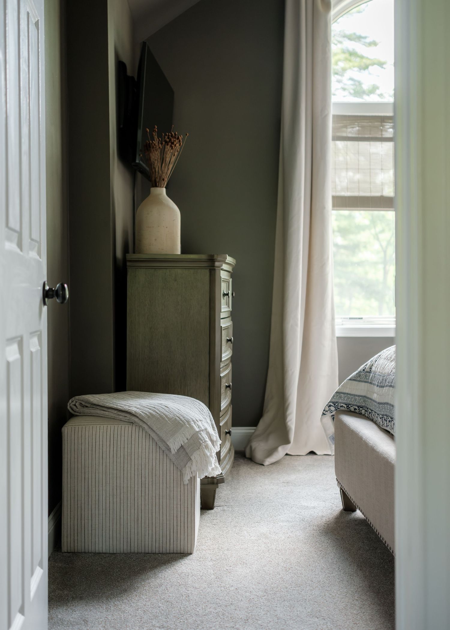 Cozy bedroom corner with olive green dresser, striped ottoman, cream curtains, and a vase of dried flowers against dark walls.