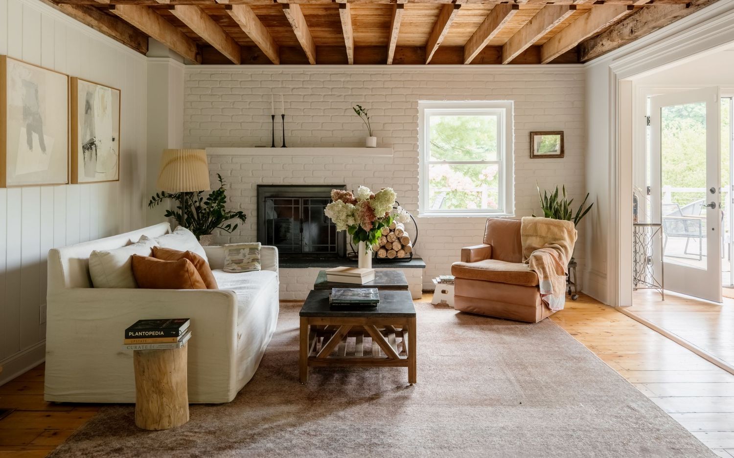 Cozy farmhouse living room with white brick fireplace, exposed wood beam ceiling, neutral sofa, and vintage coffee table.