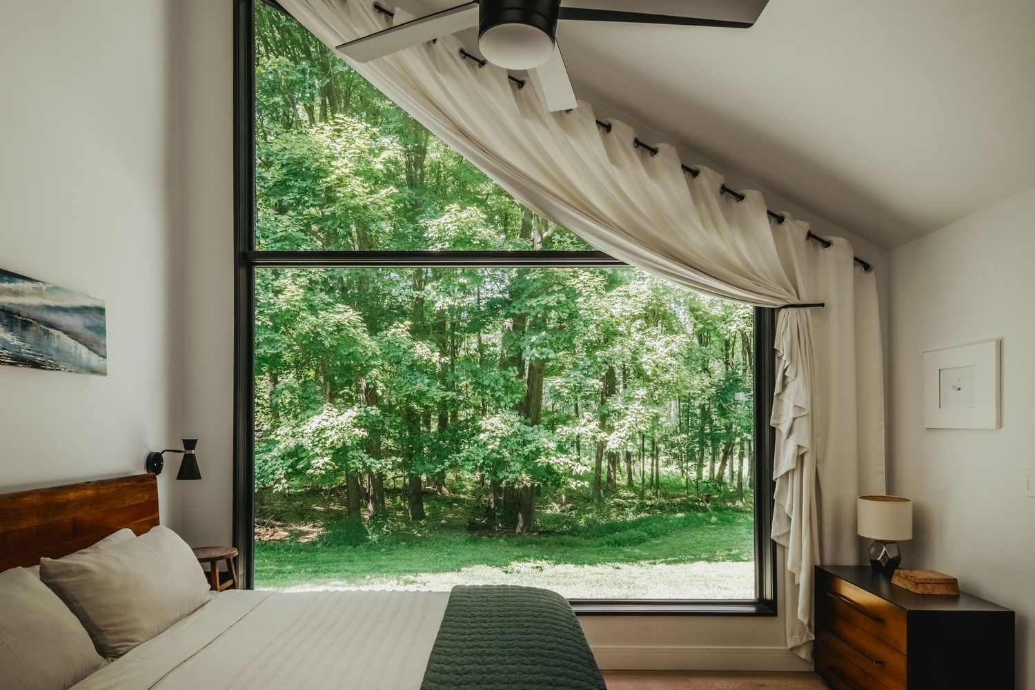 A bedroom with windows overlooking a green forest features a white bed, lamp, and linen walls.