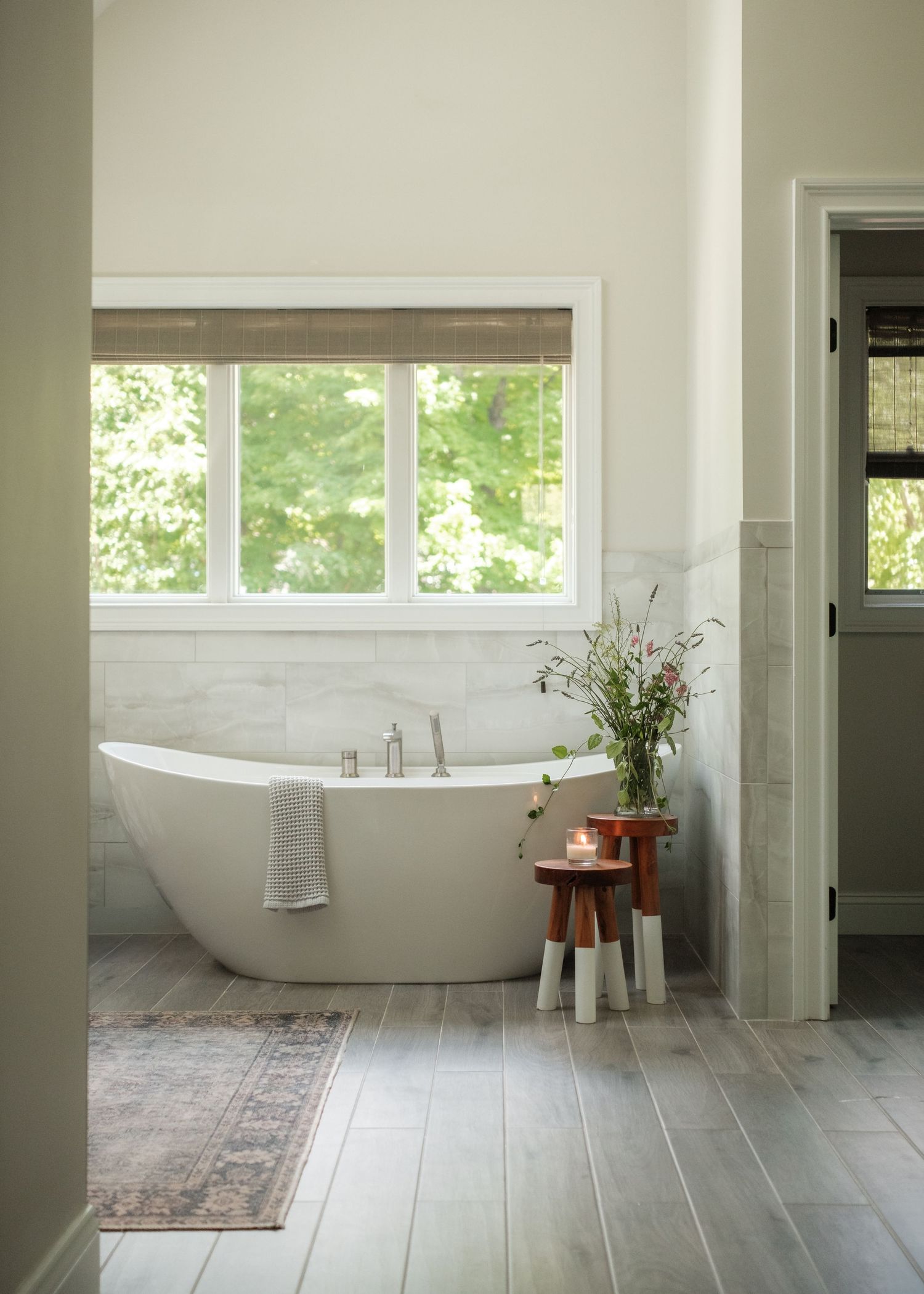 Serene bathroom with freestanding white soaking tub, marble tile wall, wood-look floors, and a window overlooking lush greenery.