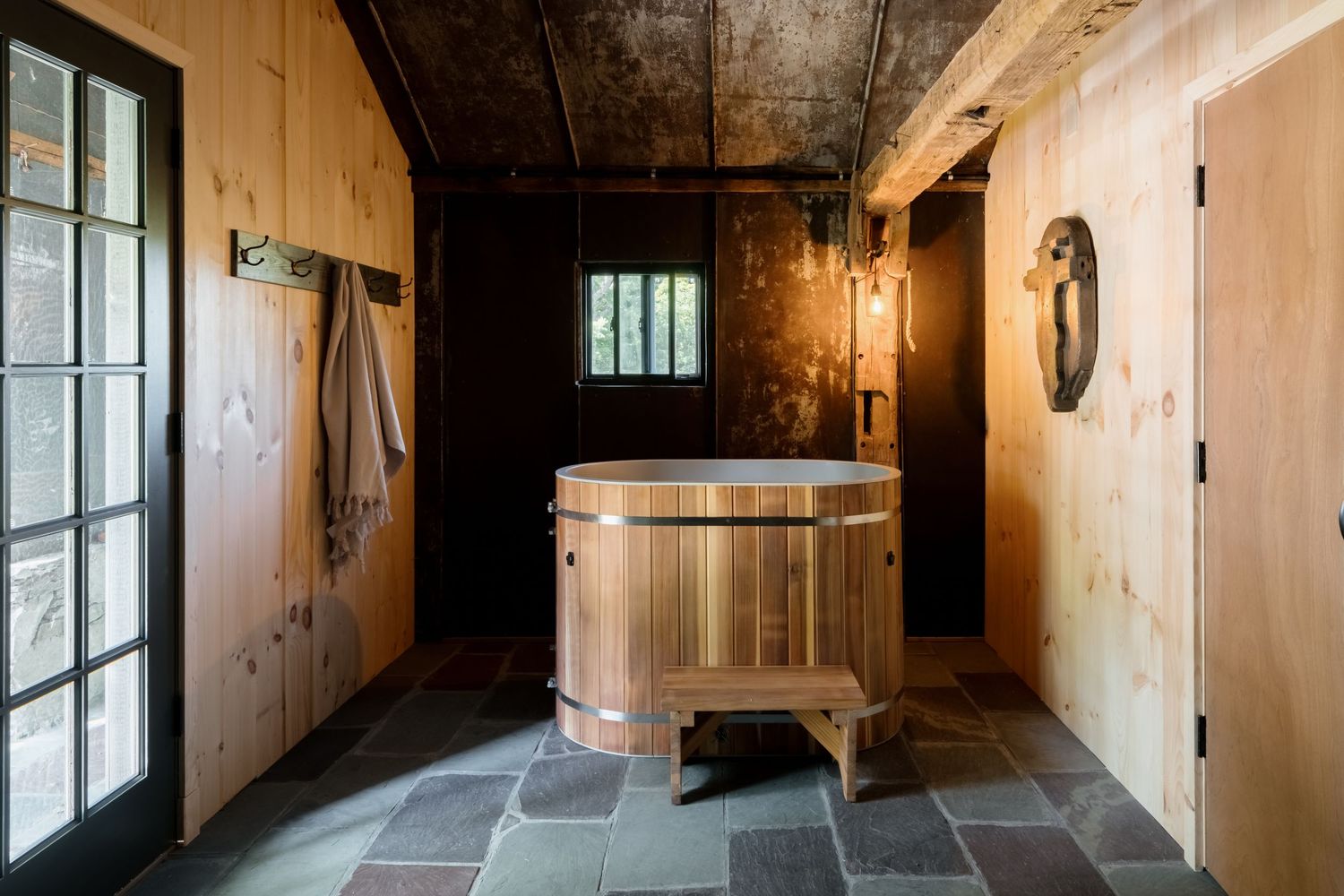 Rustic wooden bathroom with a cedar soaking tub on slate tile flooring, wood-paneled walls, and natural lighting.