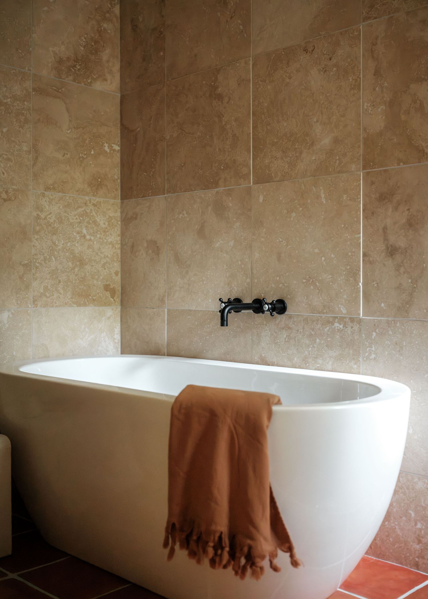 Freestanding white bathtub with terracotta towel against beige travertine tile wall with black wall-mounted faucet.
