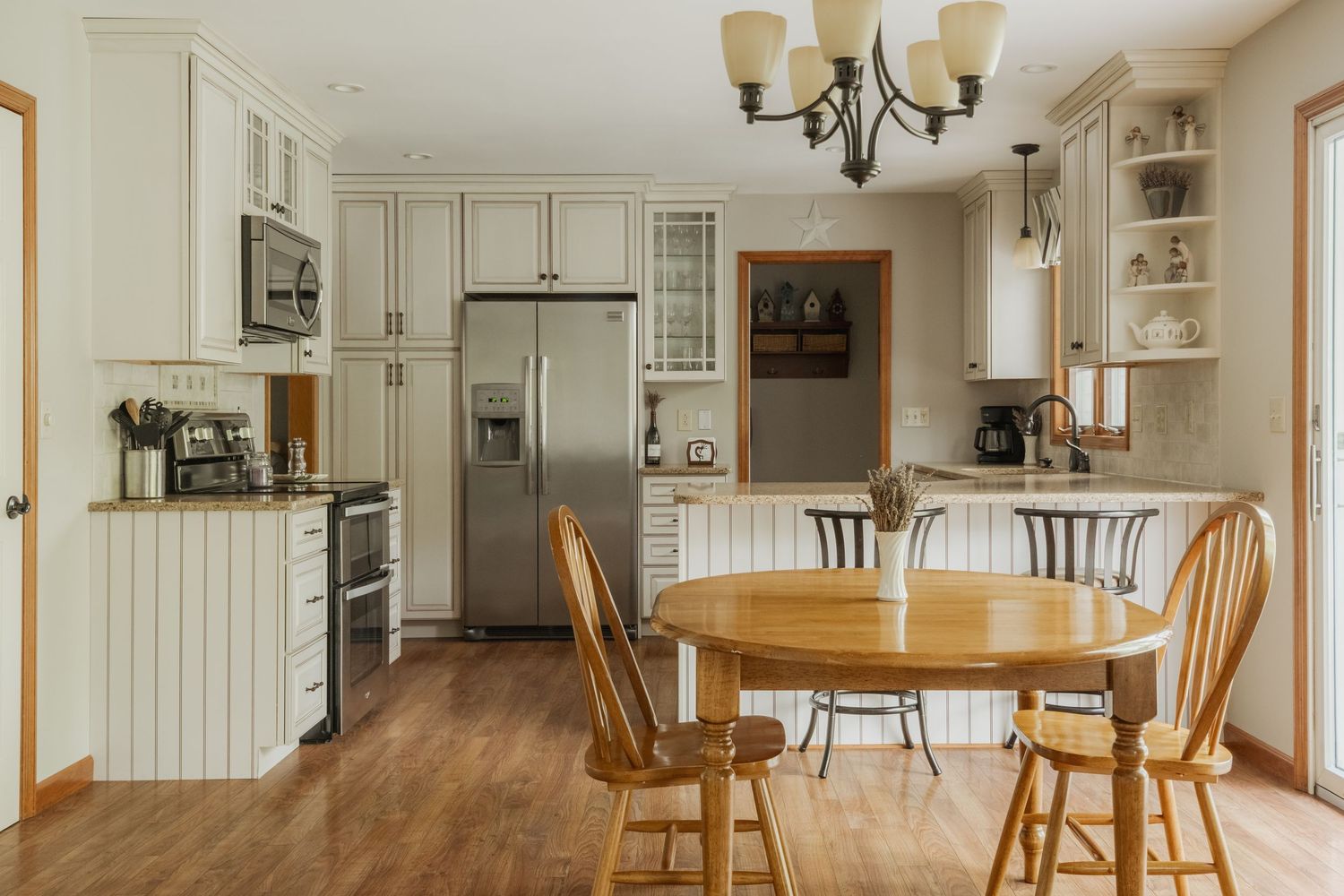 Bright kitchen with white cabinets, stainless steel fridge, wood dining table, chairs, and hardwood floors under a chandelier.
