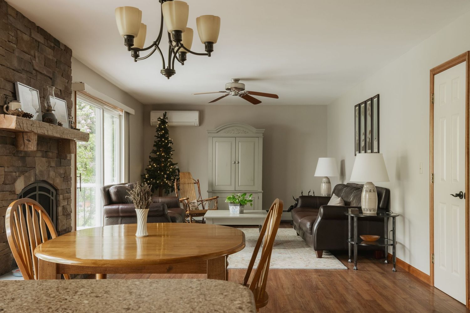 Open-plan dining and living room with stone fireplace, oval wood table, ceiling fans, and decorated Christmas tree.