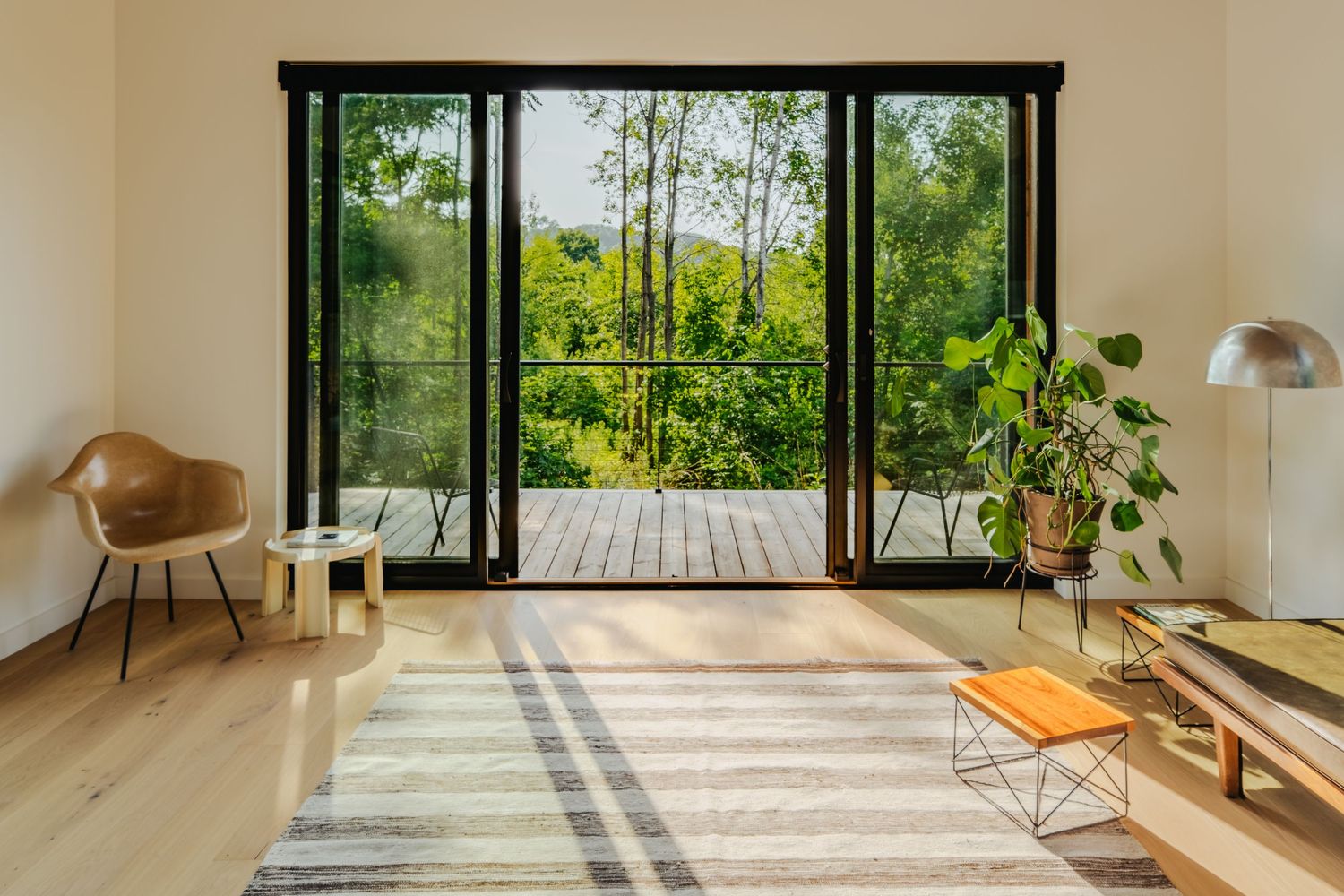 Modern minimalist living room with floor-to-ceiling windows showcasing lush green forest views and natural light.
