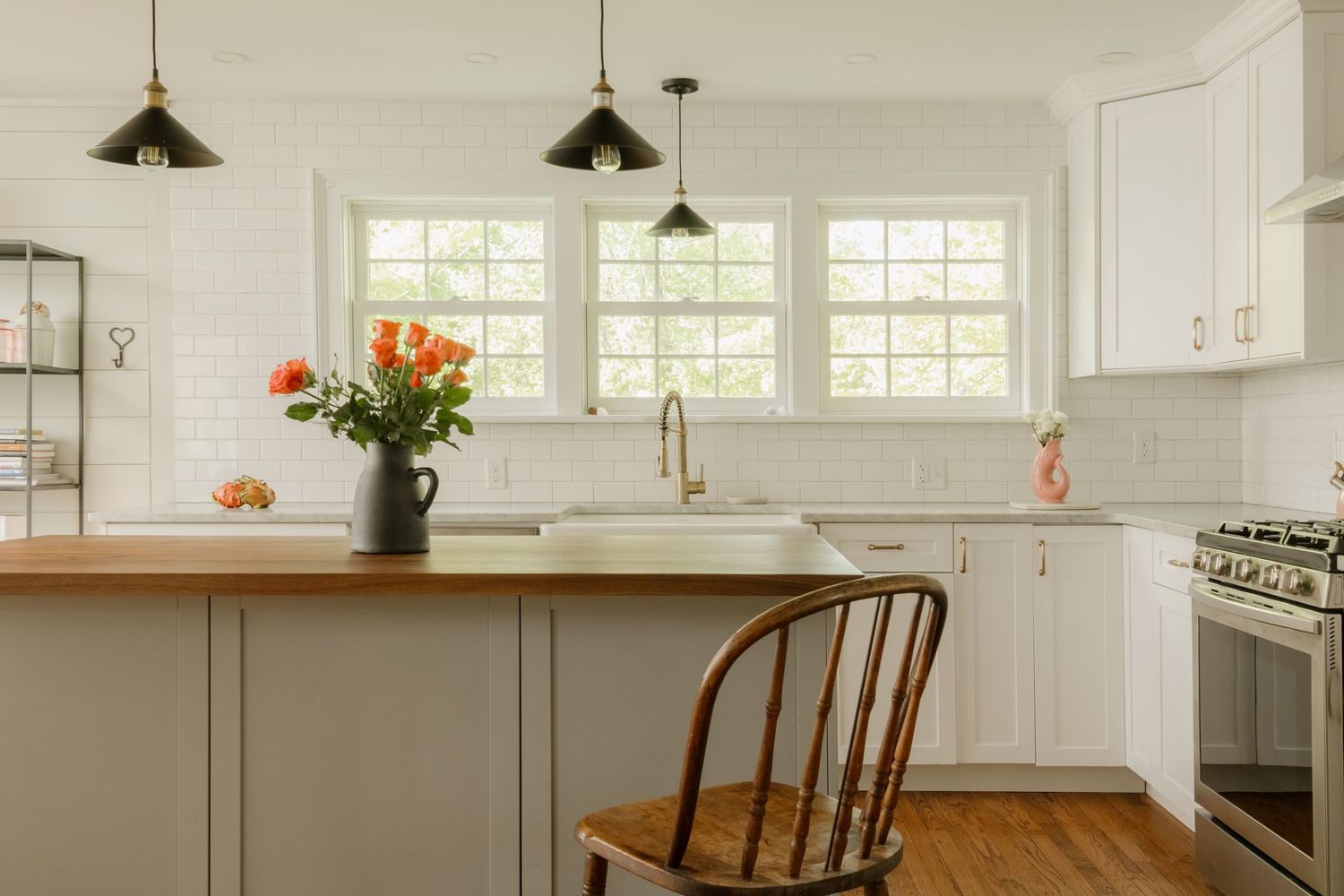White farmhouse kitchen with wood island, black pendant lights, apron sink, and orange flowers by bright windows.