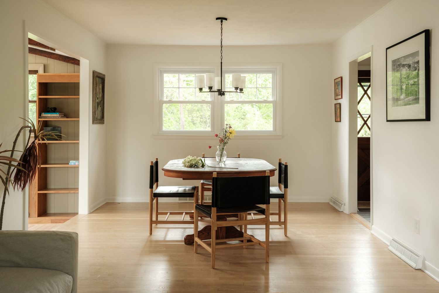 Bright dining room with round wooden table, black chairs, hardwood floors, and a chandelier near large windows.