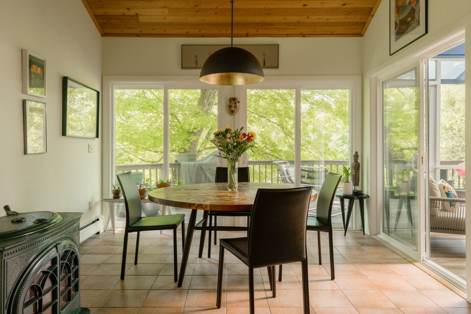Bright dining room with round wood table, green chairs, pendant light, and large windows overlooking lush trees.