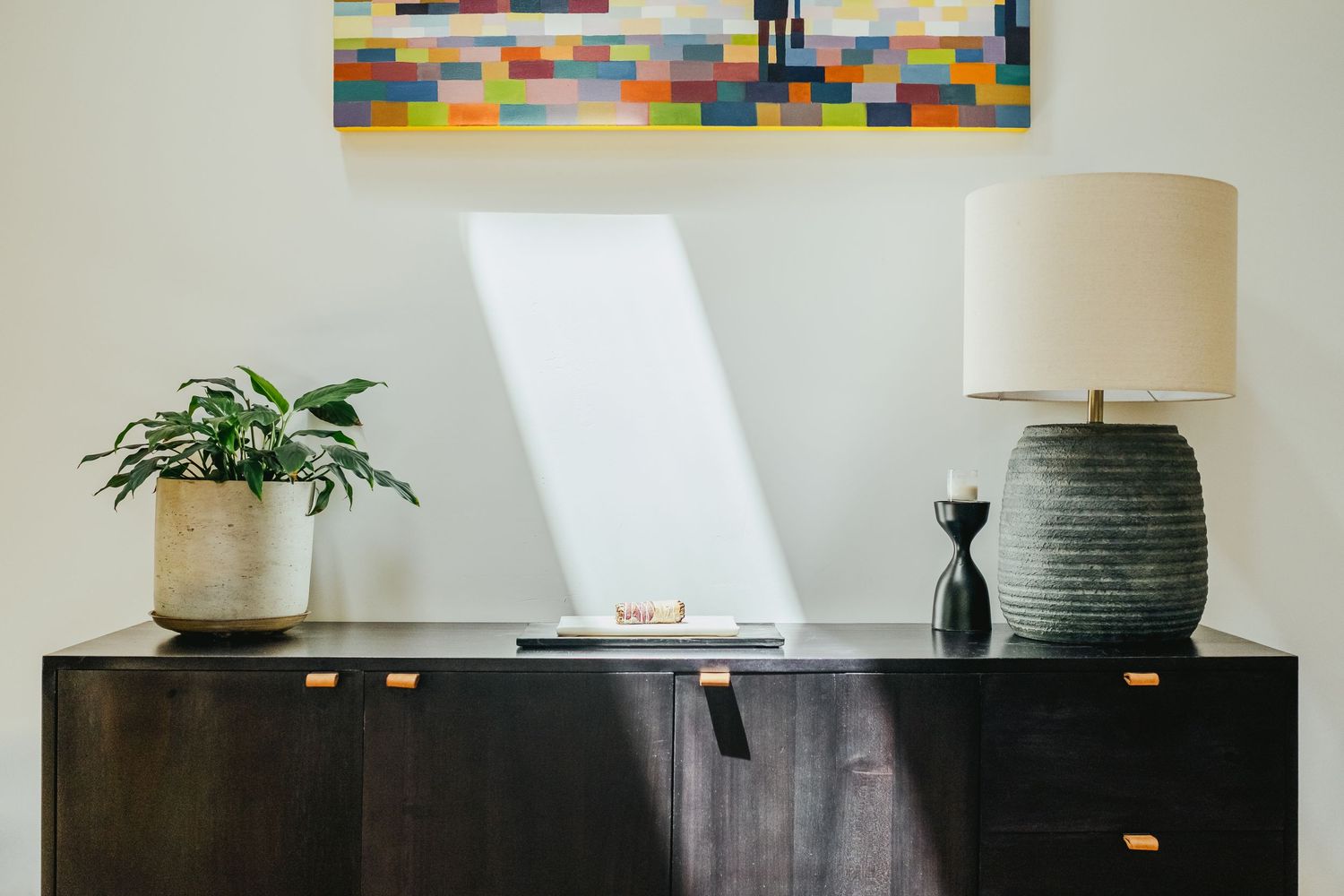 Modern black credenza styled with a textured ceramic lamp, potted plant, and colorful abstract wall art above.