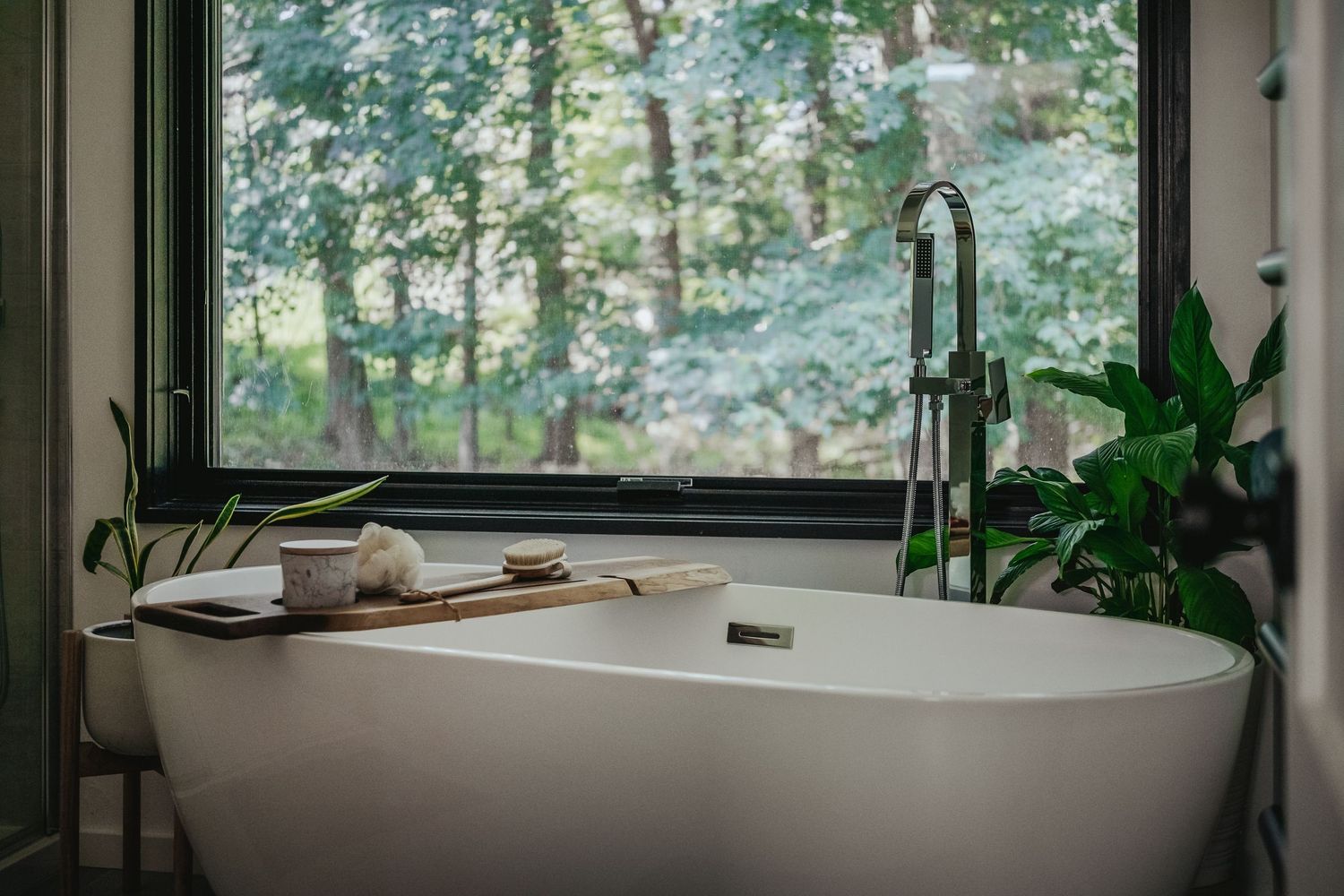 Modern freestanding white bathtub next to large window with forest view and tall black faucet fixture.