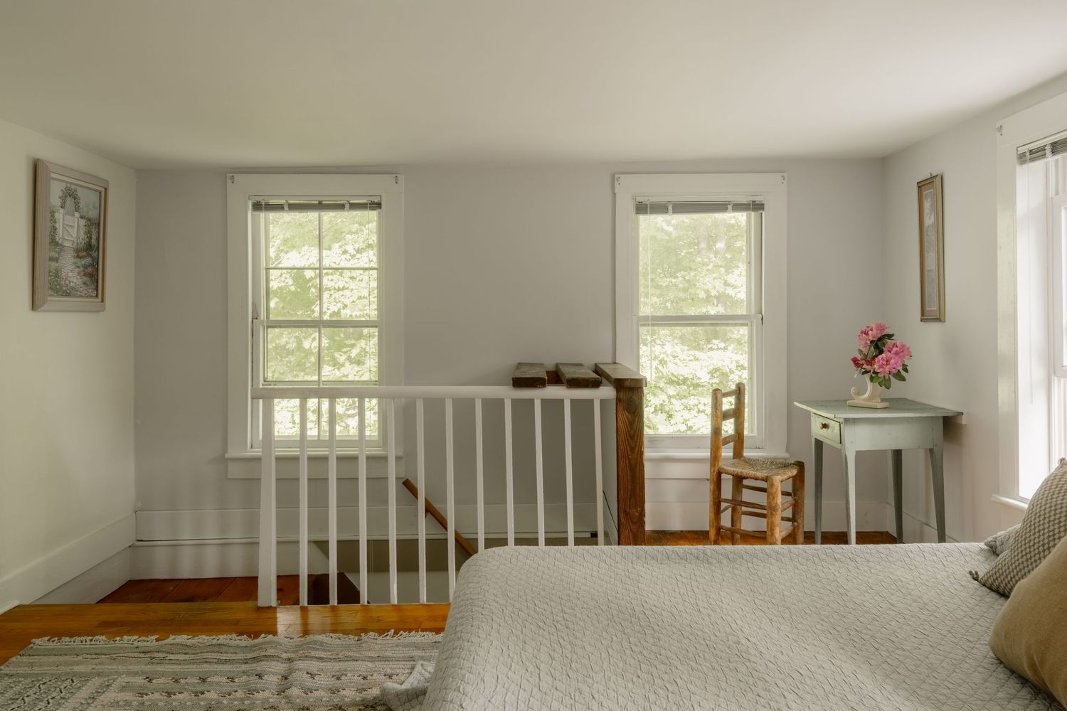 Bright bedroom with white railings, hardwood floors, two windows, a wooden chair, and a small blue desk with pink flowers.