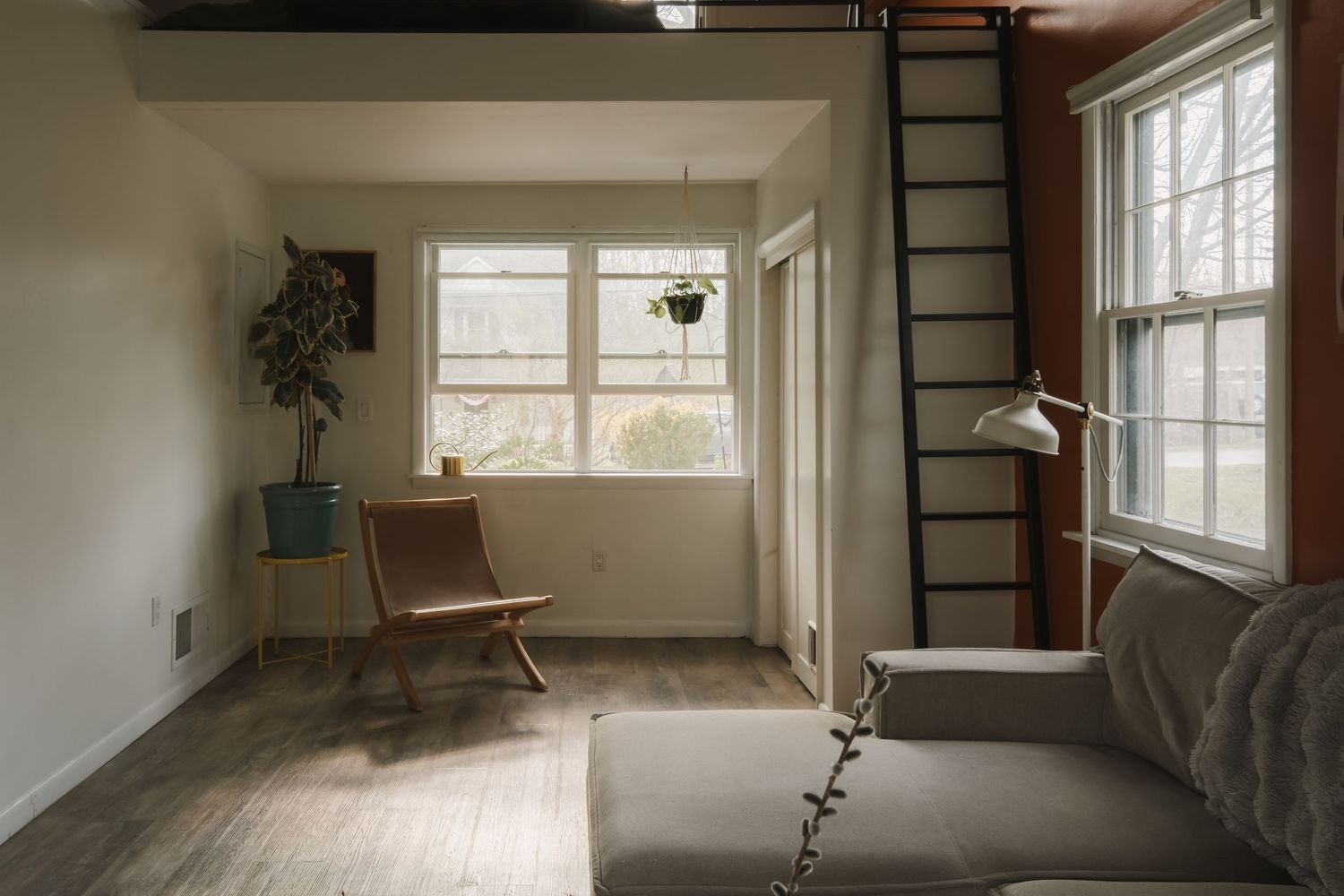 Cozy minimalist living room with wood floors, gray sofa, ladder, chair, and plants near bright windows.