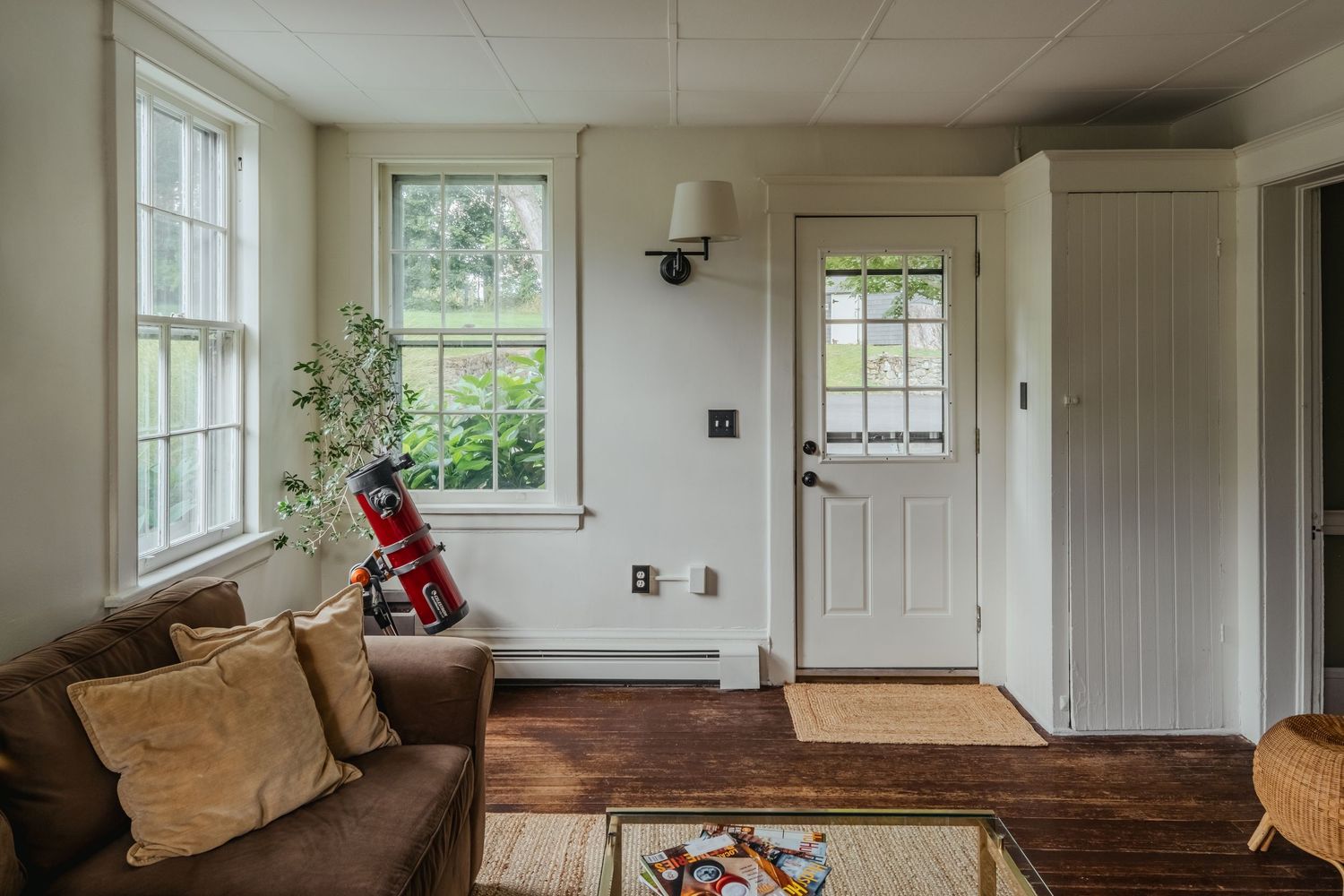 Cozy living room interior with large windows, brown couch, and white door showcasing rustic hardwood floors and natural light.