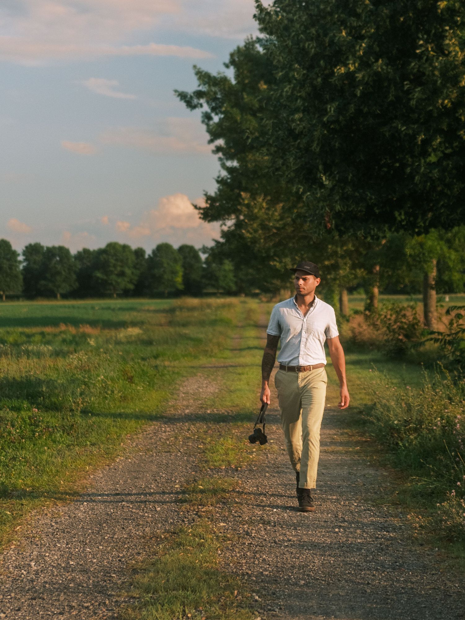 Figure in white shirt and khaki pants walks along a gravel path through a scenic countryside at golden hour.
