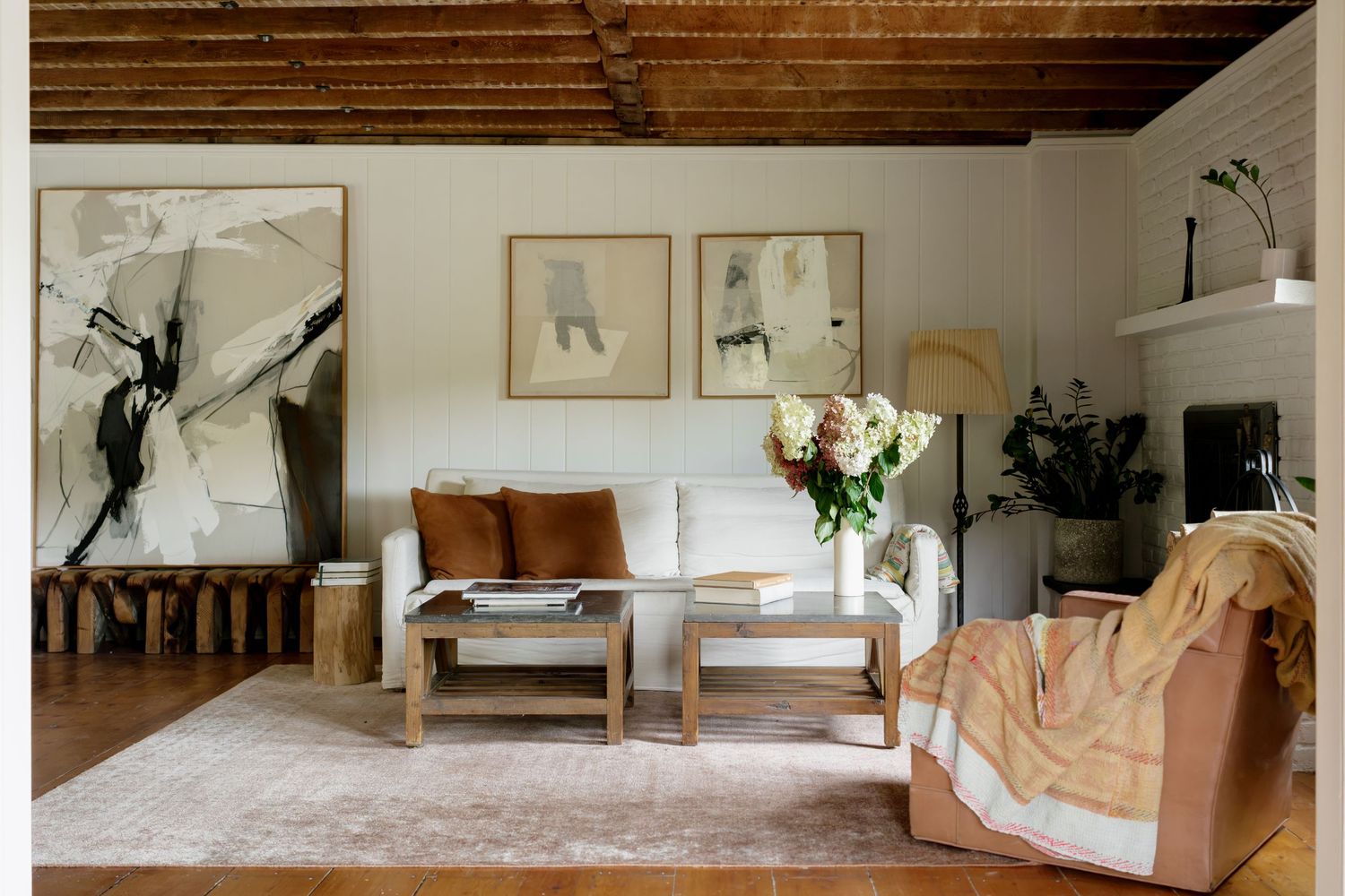 Cozy living room with white sofa, wooden ceiling, abstract art, floral arrangement, and warm neutral tones.