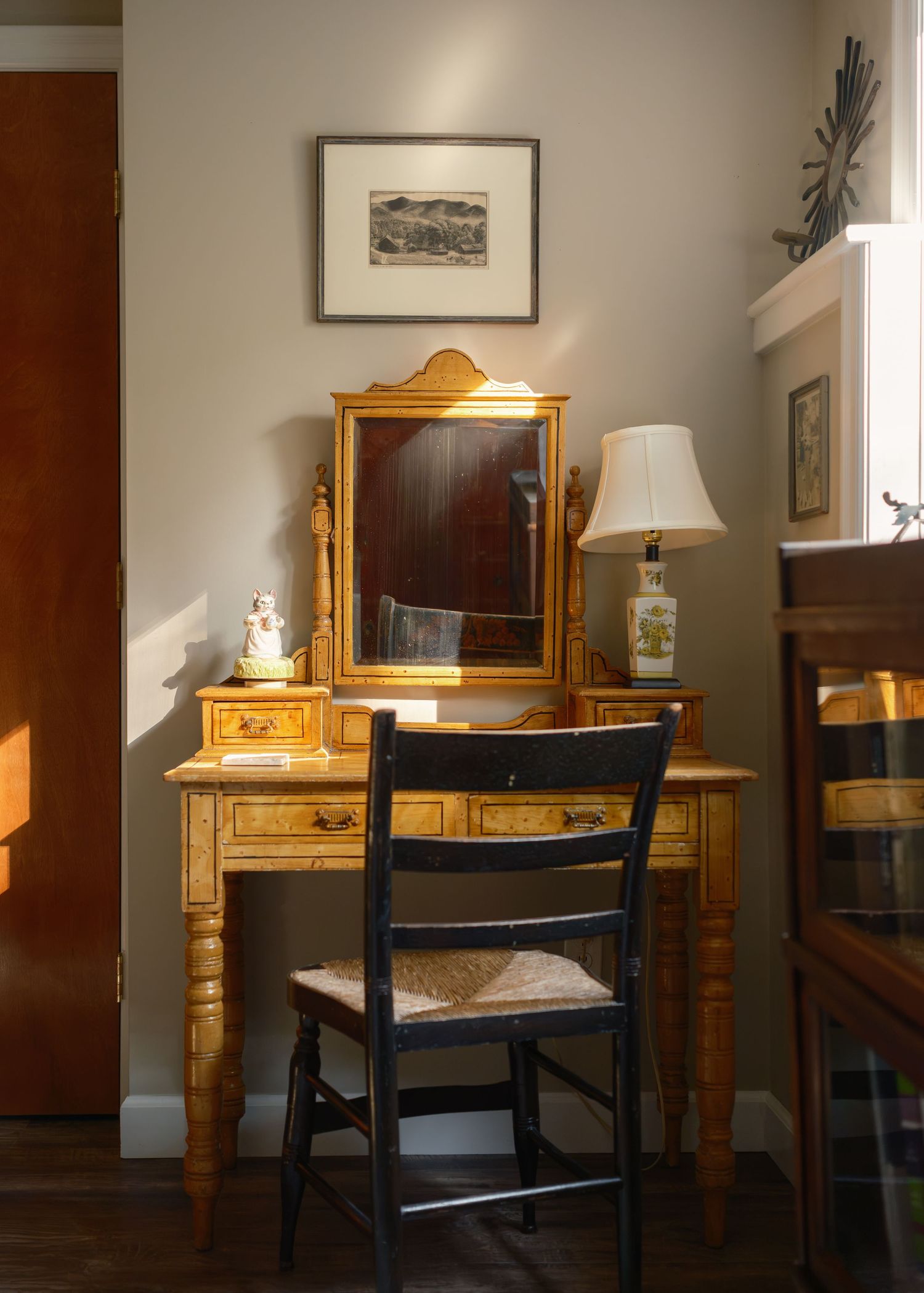 A vintage wooden vanity with mirror and black chair sits in warm golden light beside a framed wall print.