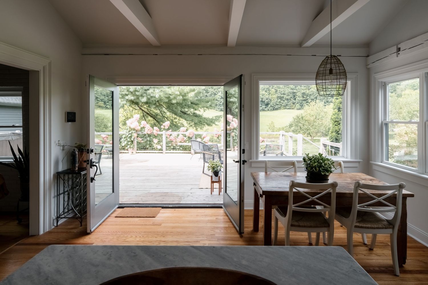 Bright dining room with open French doors leading to a sunny deck, wood table with white chairs, hardwood floors and a woven pendant light.