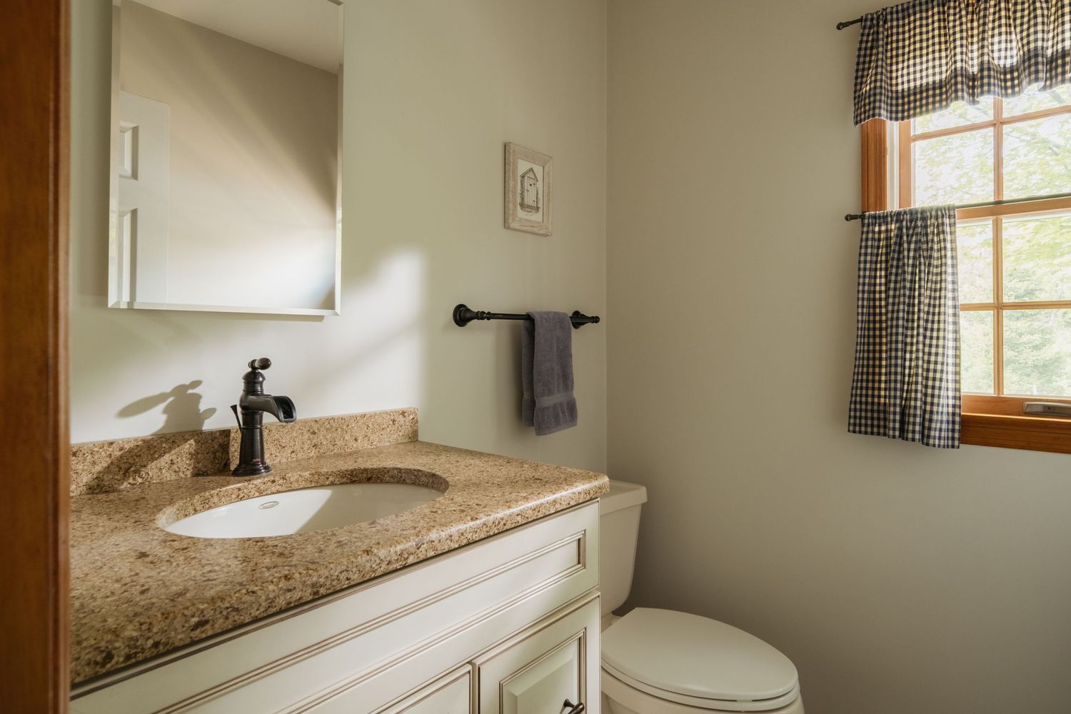A clean bathroom featuring a granite vanity with white cabinets, mirror, toilet, and a window with checkered curtains.