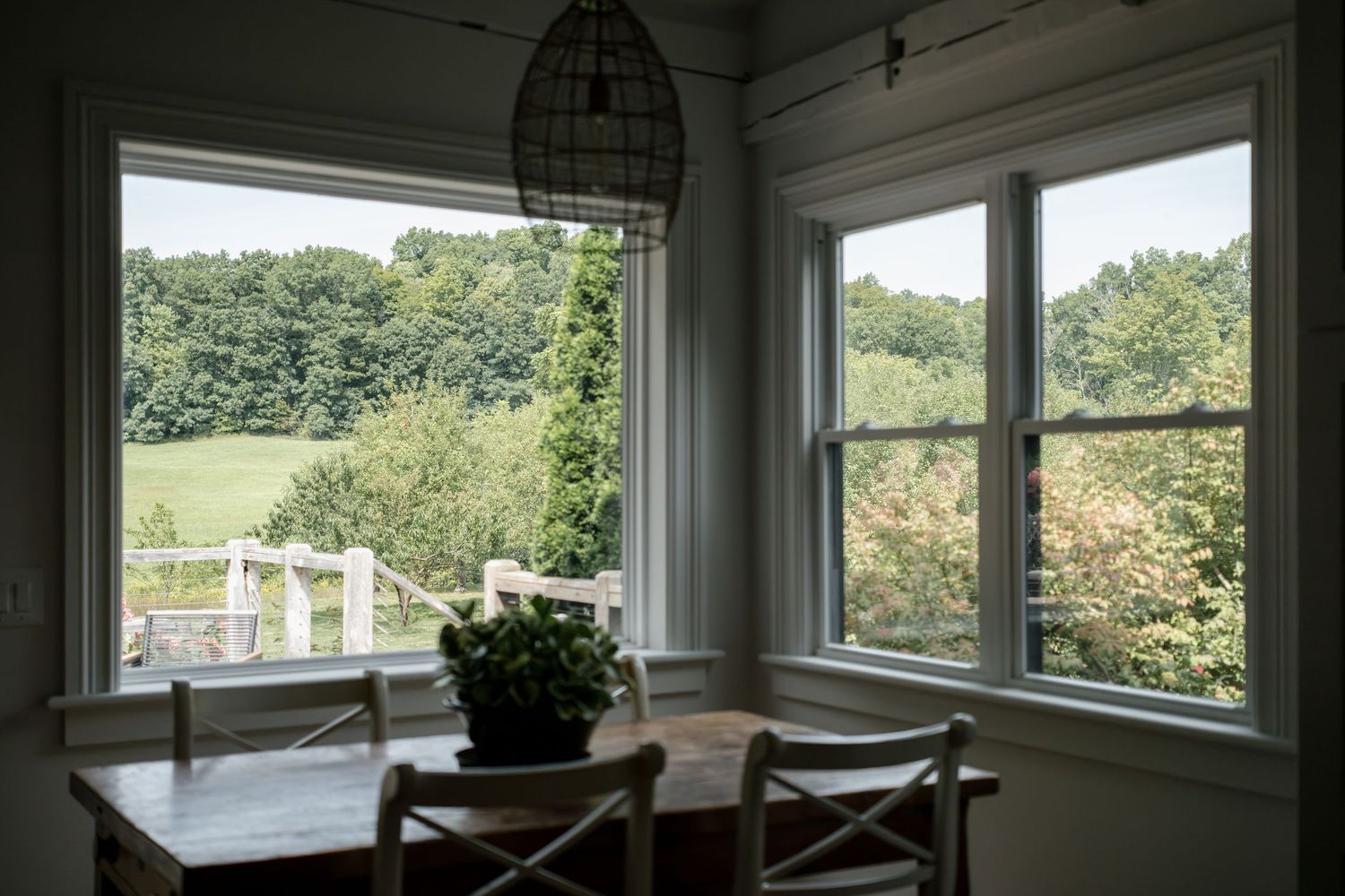 Sunlit farmhouse dining nook with wooden table, cross-back chairs, potted plant and windows overlooking green fields and trees.