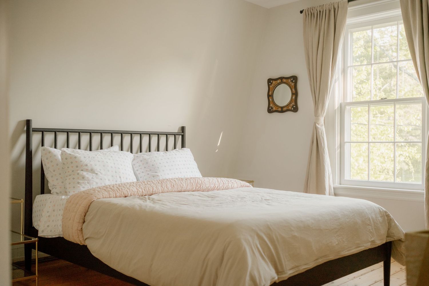 Cozy bedroom with black metal bed frame, white and pink bedding, hardwood floors, and a bright window with curtains.