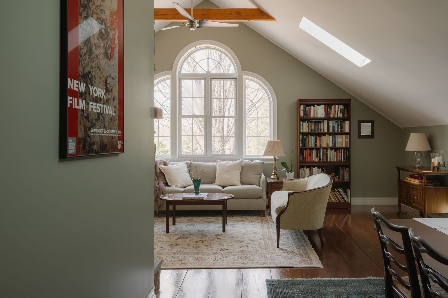Cozy living room with arched window, skylight, bookshelf, sofa, and hardwood floors in a sloped ceiling space.