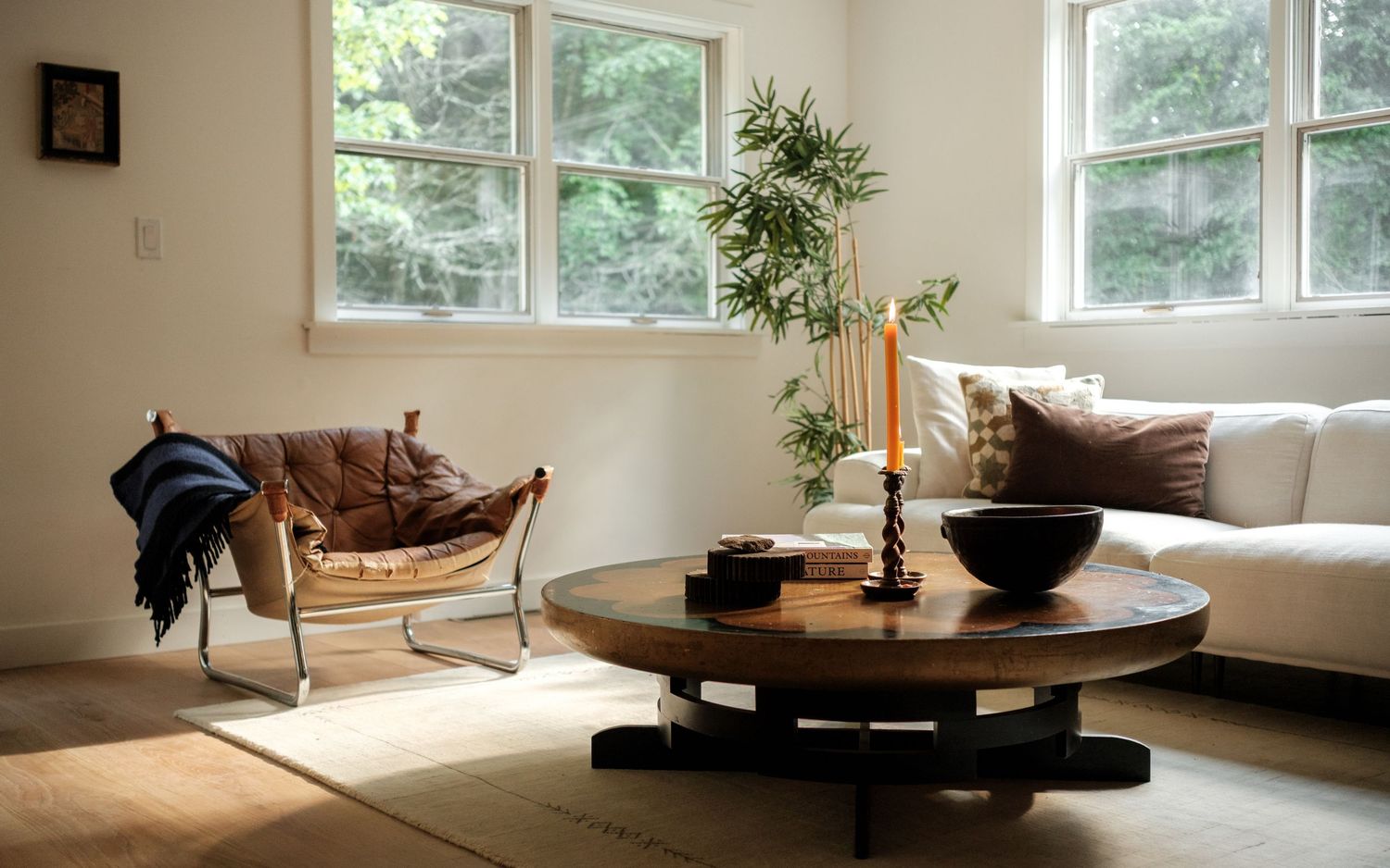 Cozy minimalist living room with round wooden coffee table, white sofa, bamboo plant, and natural light from large windows.