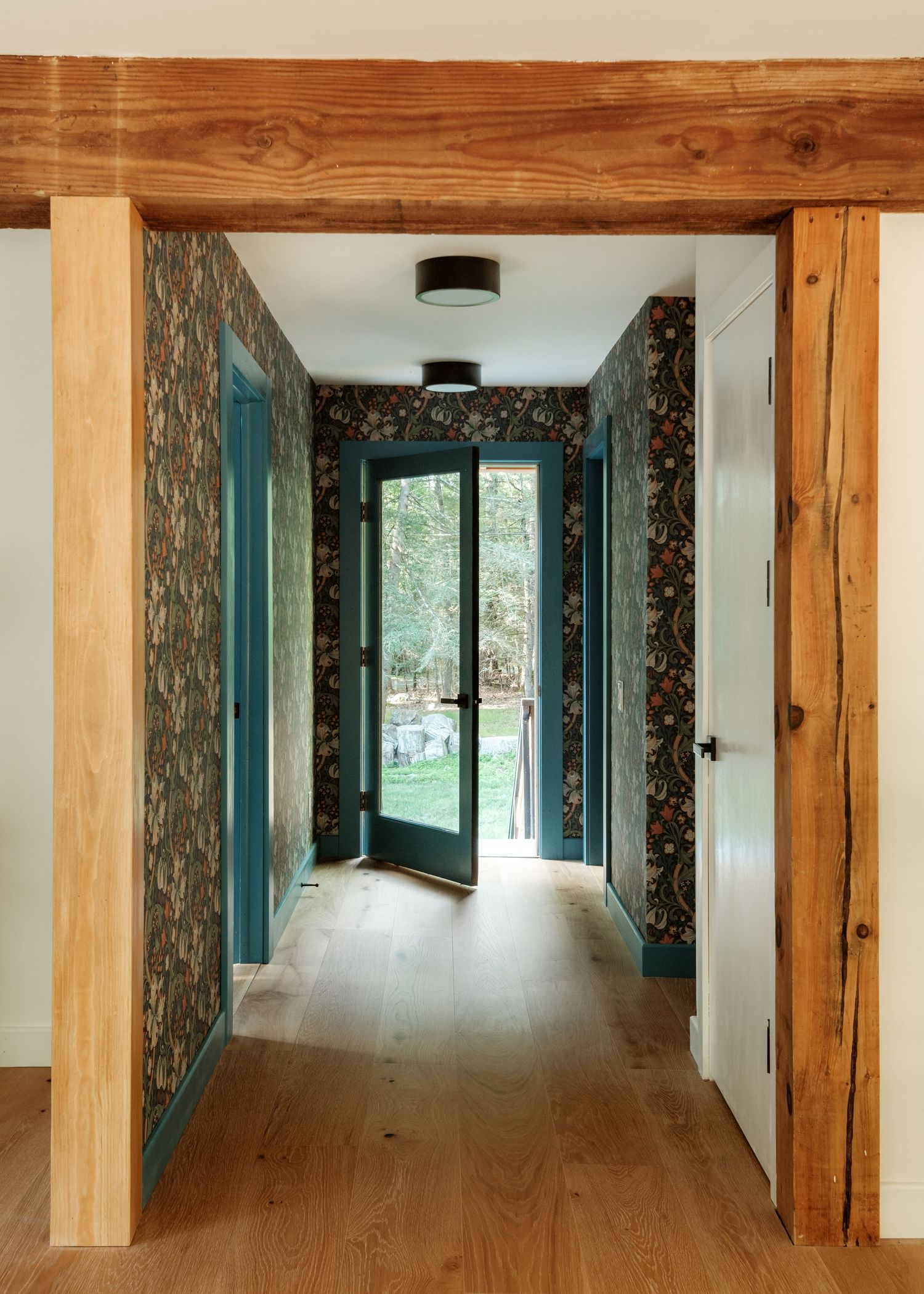 Hallway with floral wallpaper, teal door trim, hardwood floors, and rustic wood beam framing leading to glass exterior doors.