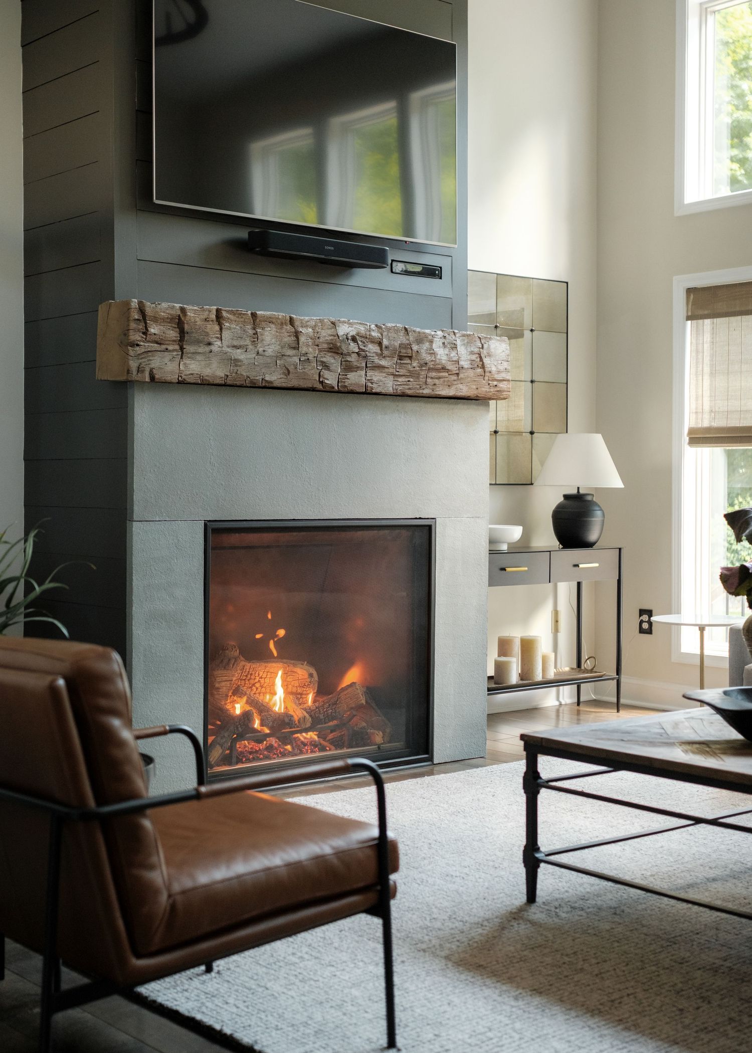 Modern living room with lit fireplace, rustic wood mantel, leather armchair, and wall-mounted TV above stone surround.