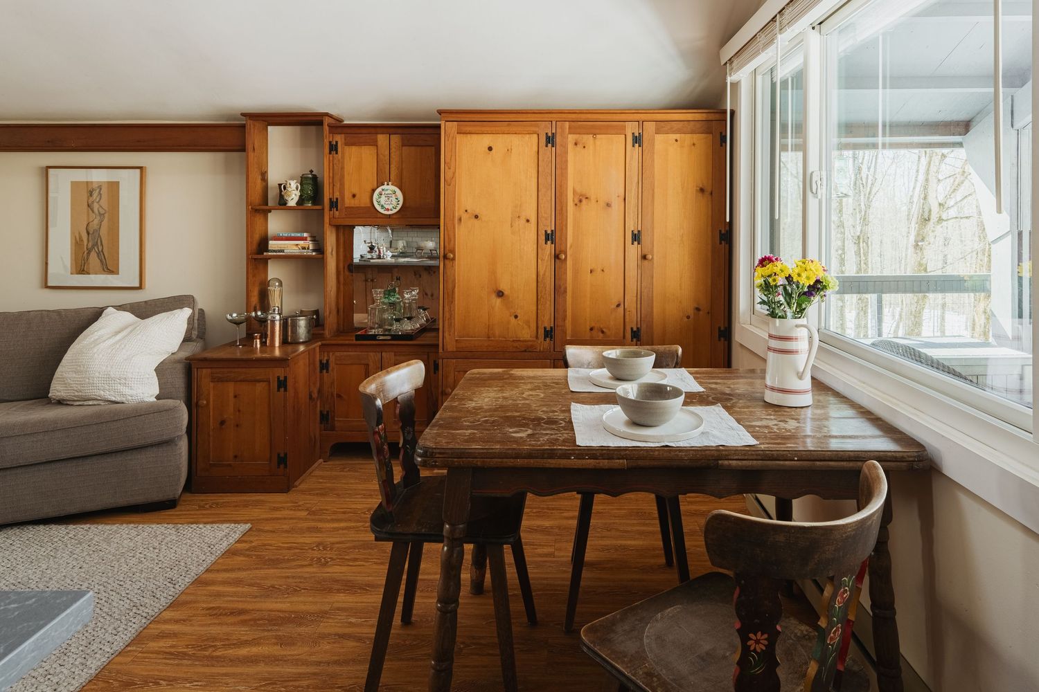 Rustic wood-paneled dining room with large windows, natural light, and modern furnishings creates a cozy farmhouse atmosphere.