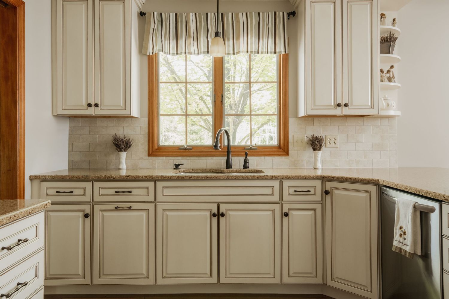 Cream kitchen cabinets with granite countertops, a farmhouse sink, and a striped valance framing a wood-trimmed window.