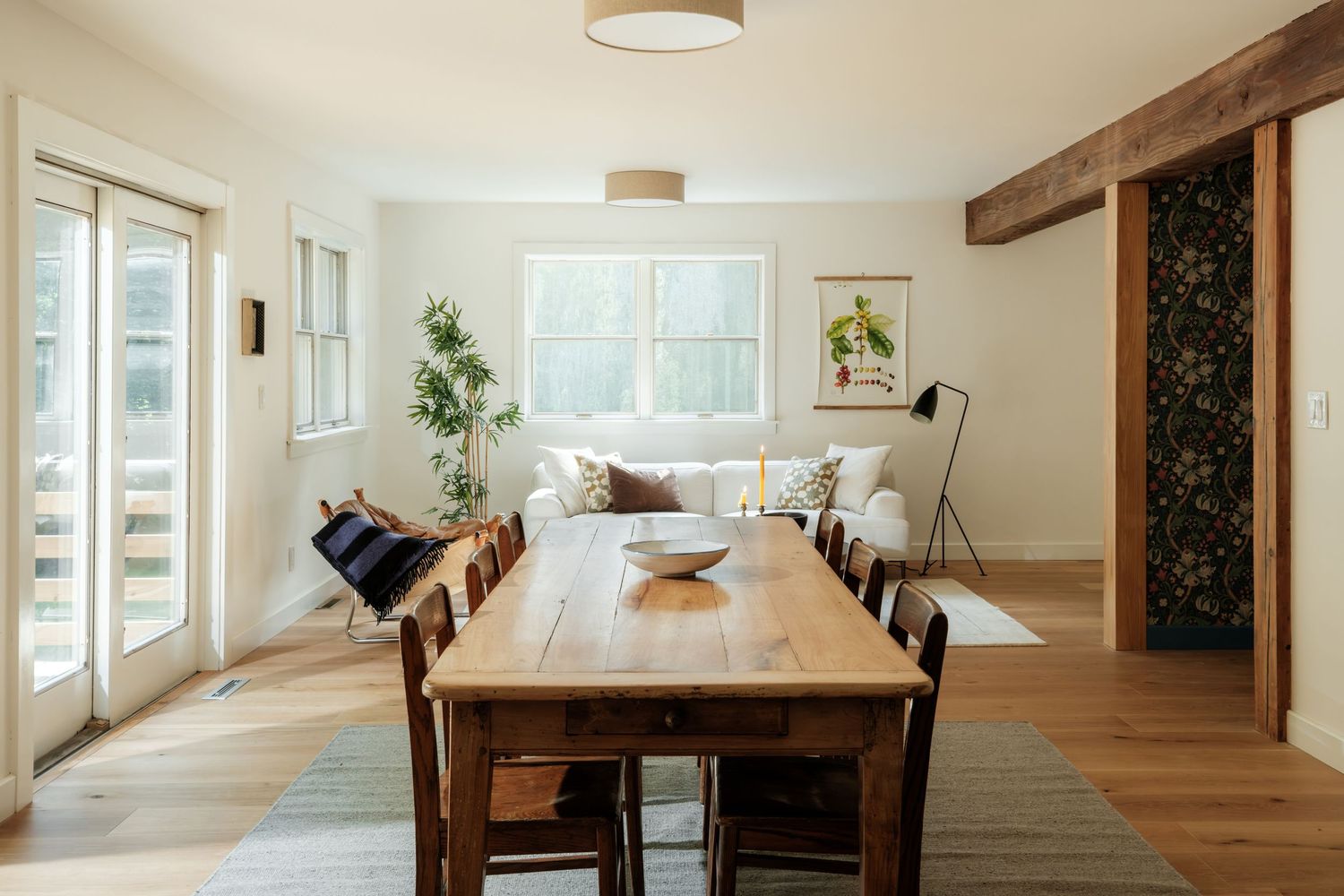 Bright dining room with rustic wood table, chairs, white sofa, exposed beam, and hardwood floors in a modern farmhouse.