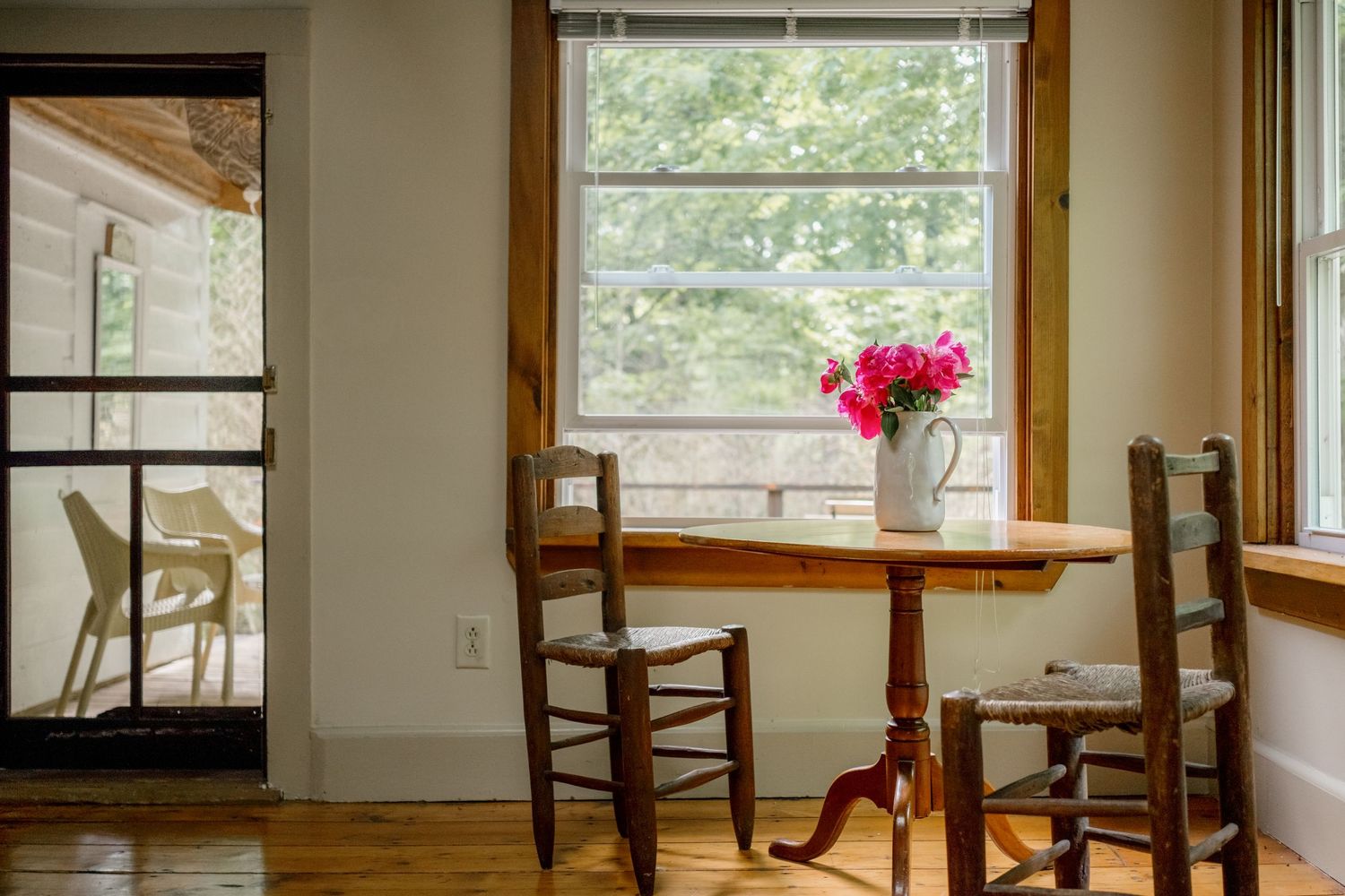 Cozy dining nook with wooden table, ladder-back chairs, pink flowers, and bright window overlooking lush greenery.