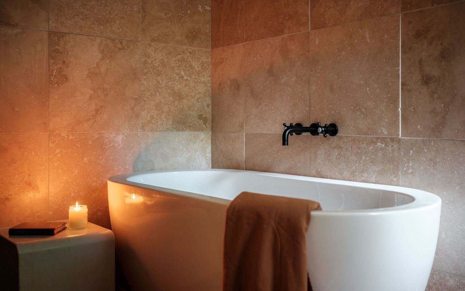 Elegant freestanding white bathtub with black wall-mounted faucet, warm stone tiles, candles, and a folded towel.