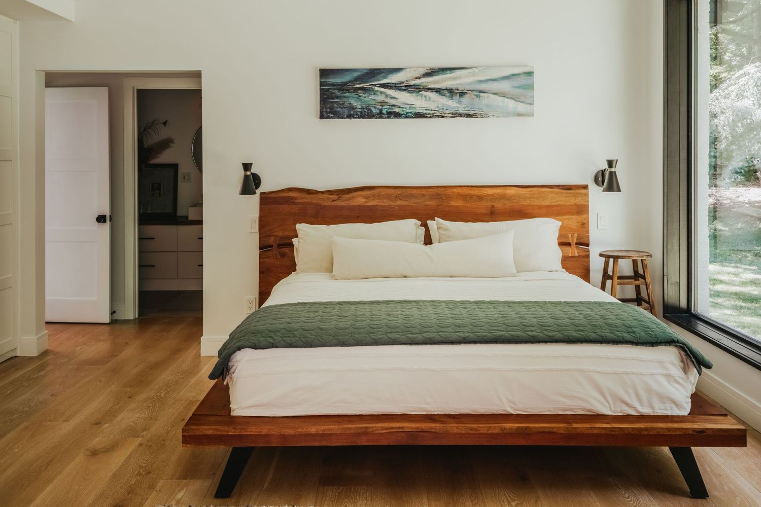 Modern bedroom with wooden platform bed, white bedding, olive green throw, and minimalist coastal artwork above headboard.
