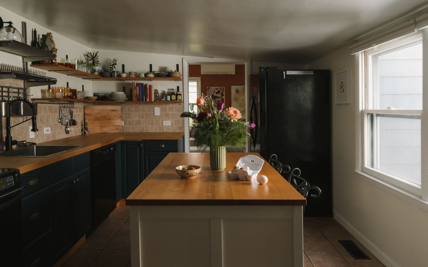 Modern farmhouse kitchen with navy cabinets, butcher block island, open shelving, and fresh flowers in natural light.