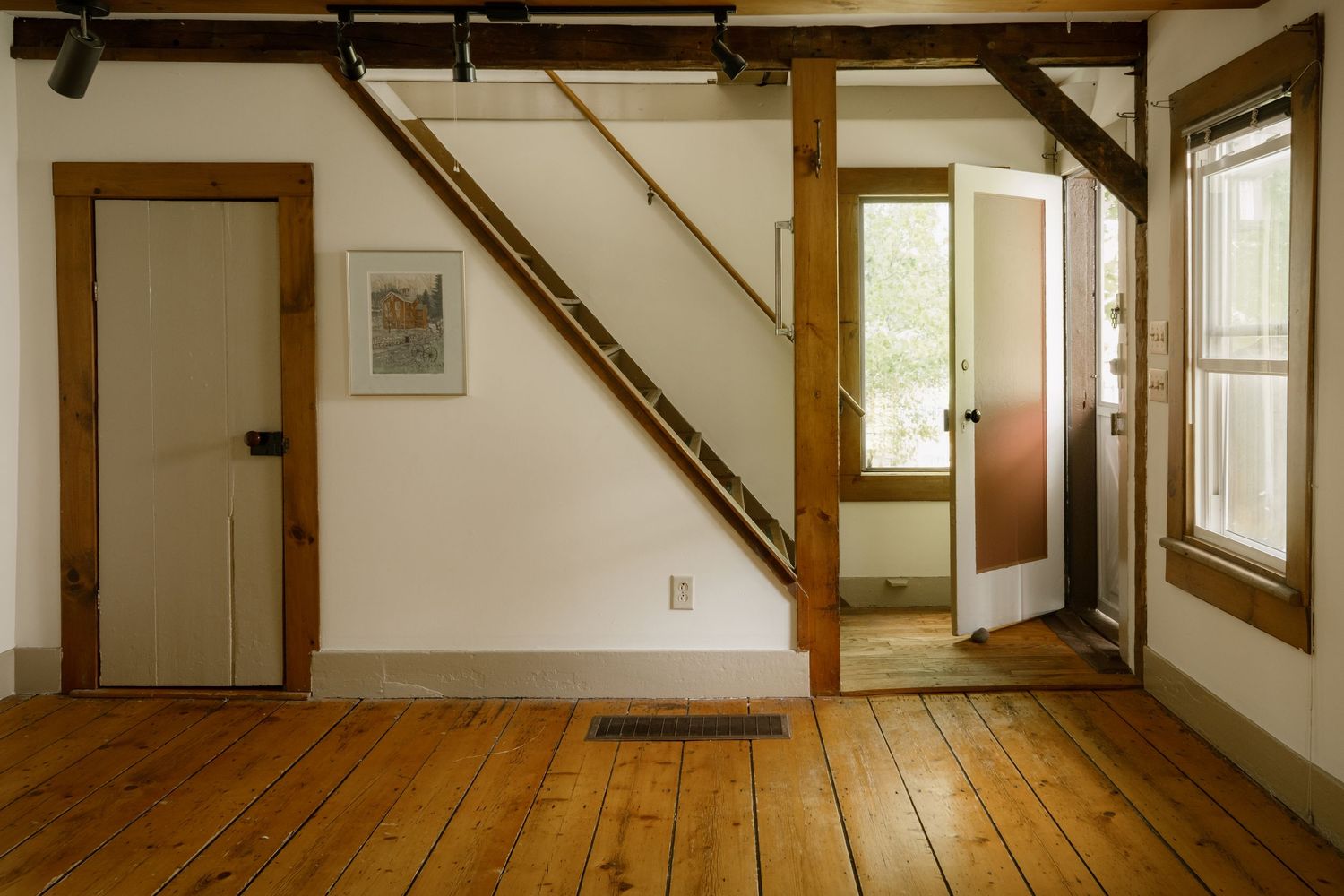 Rustic interior with hardwood floors, wooden beams, white walls, staircase, and natural light through glass door.