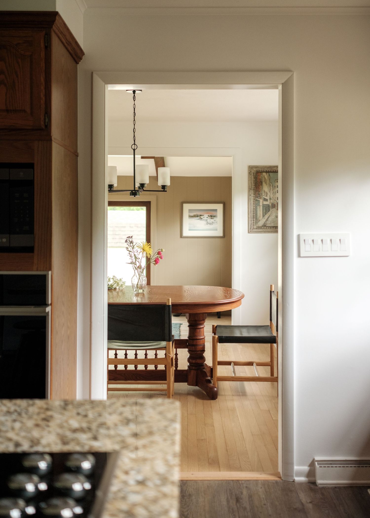 View through doorway into dining room with round wood pedestal table, black chairs, chandelier, and hardwood floors.