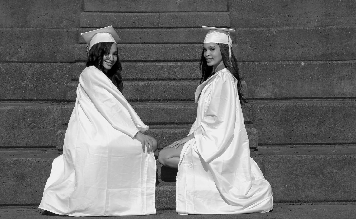 Two graduates in white gowns sitting on stone steps in black and white.
