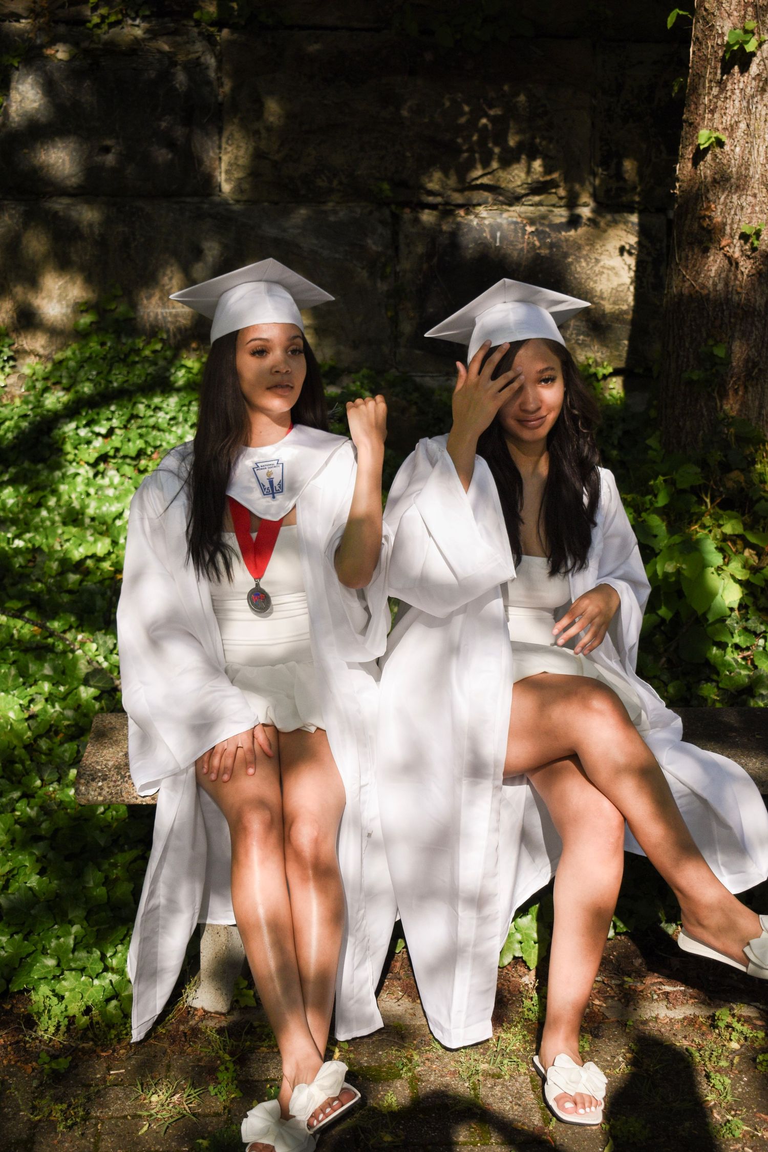 Two graduates in white gowns sitting together on a stone wall surrounded by greenery.