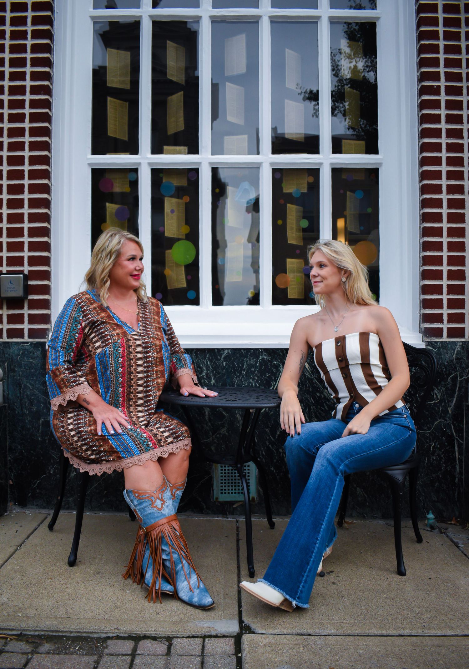 Two people in contrasting outfits sitting on chairs outside a historic building with paned windows.