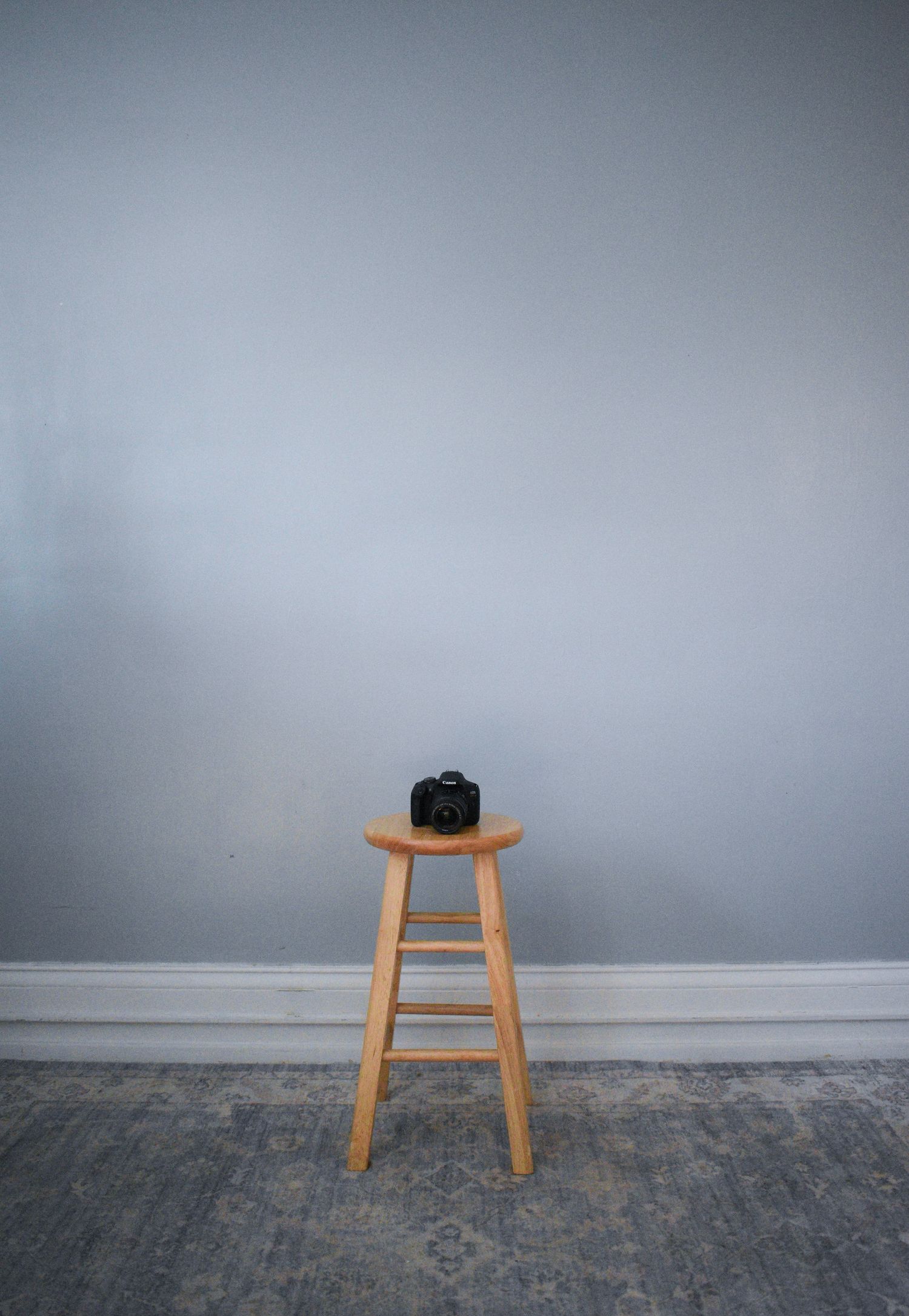 A black DSLR camera sits alone on a wooden stool against a plain grey wall.