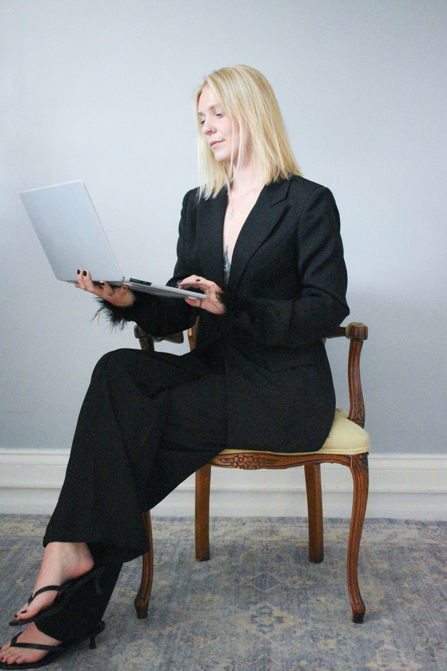 Casual business poses with a laptop on an elegant upholstered chair against gray backdrop.